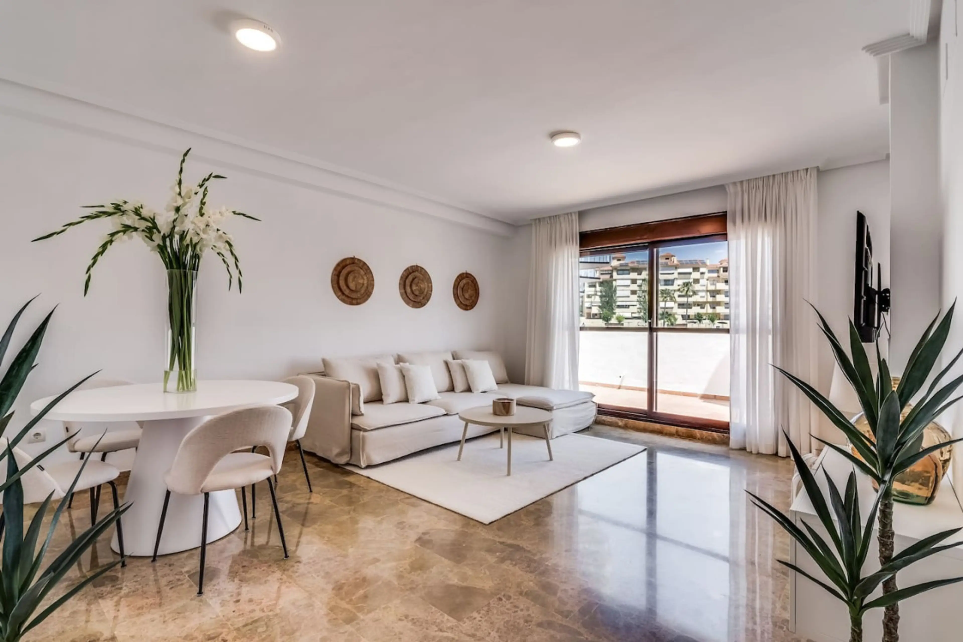 - Bright open living room with sofa and dining space ideal for family stays - Sunlit interior with balcony access modern decor and smart TV setup - Spacious holiday apartment near beach pools dining and local attractions
