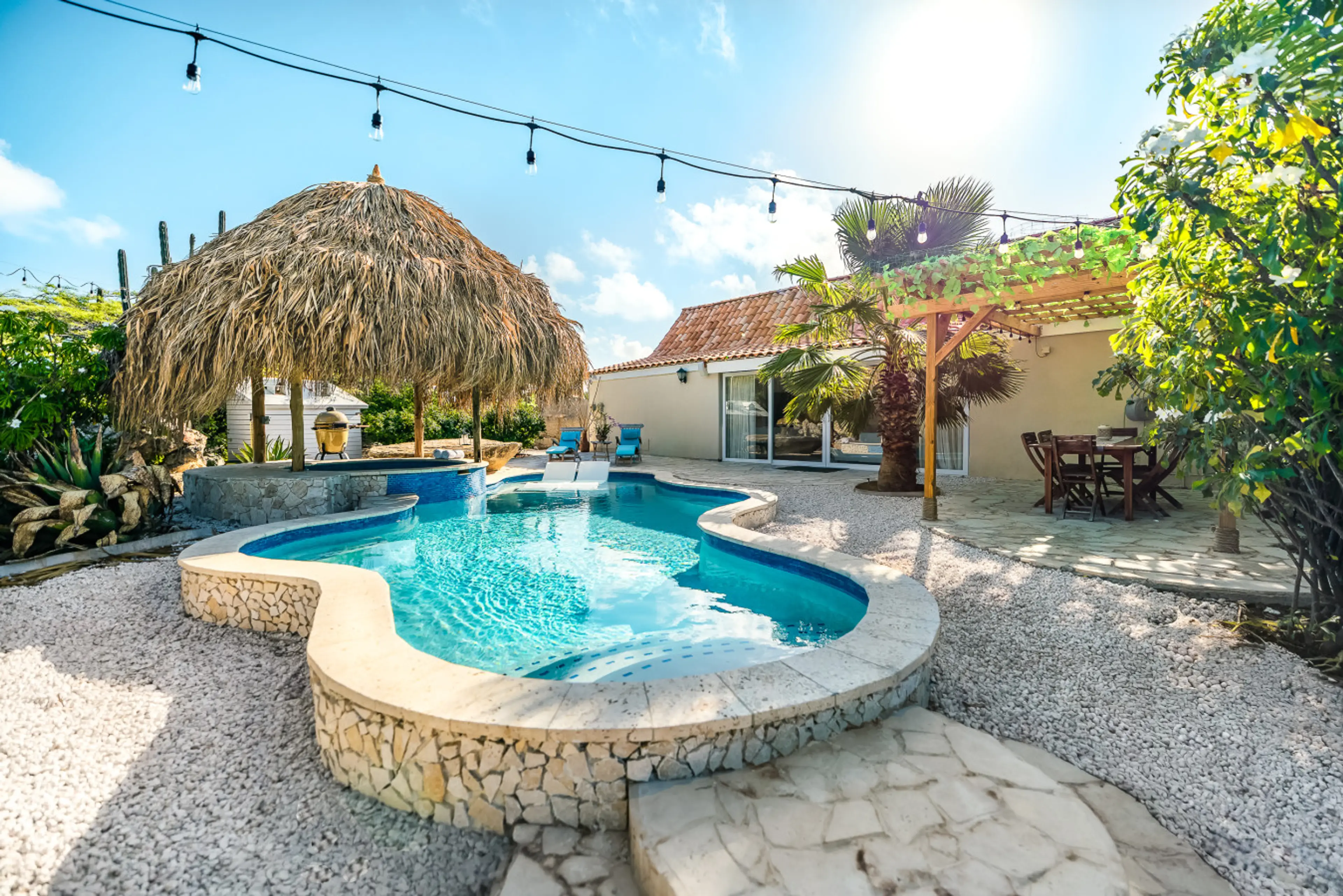 Custom pool & jacuzzi (non-heated) with 2 in-pool loungers surrounded by tropical landscaping, a shaded palapa lounge, and string lights creating a relaxed island atmosphere perfect for daytime swims and evening relaxation.