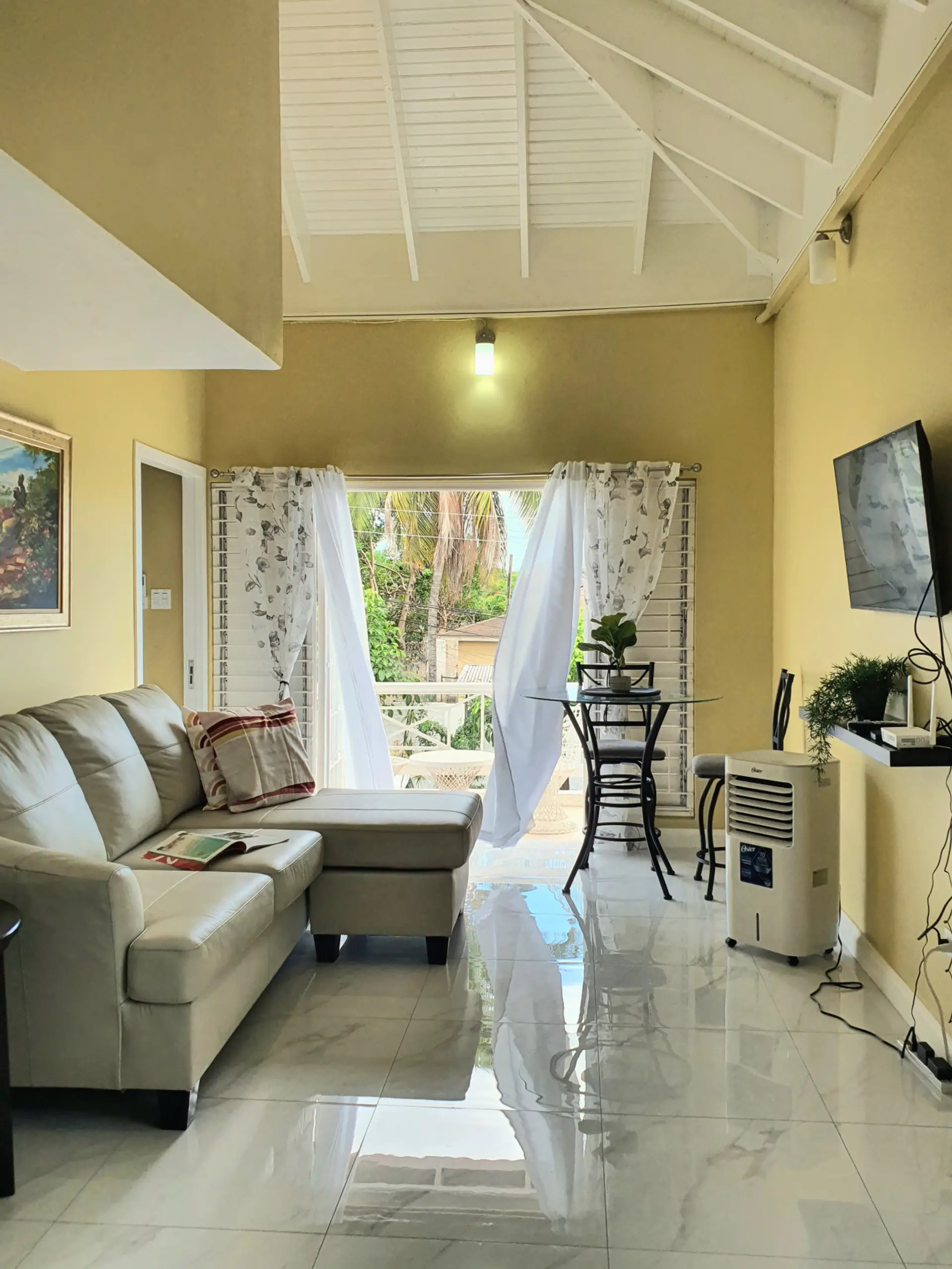 Bright living area w/ sofa & TV, opening to a breezy balcony through sheer curtains.