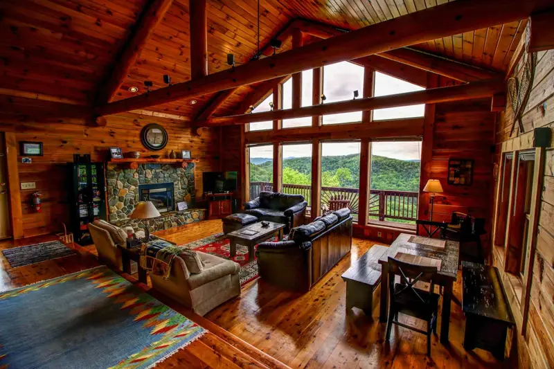 Great room with heavy timber beams, a stone fireplace, and a wall of windows