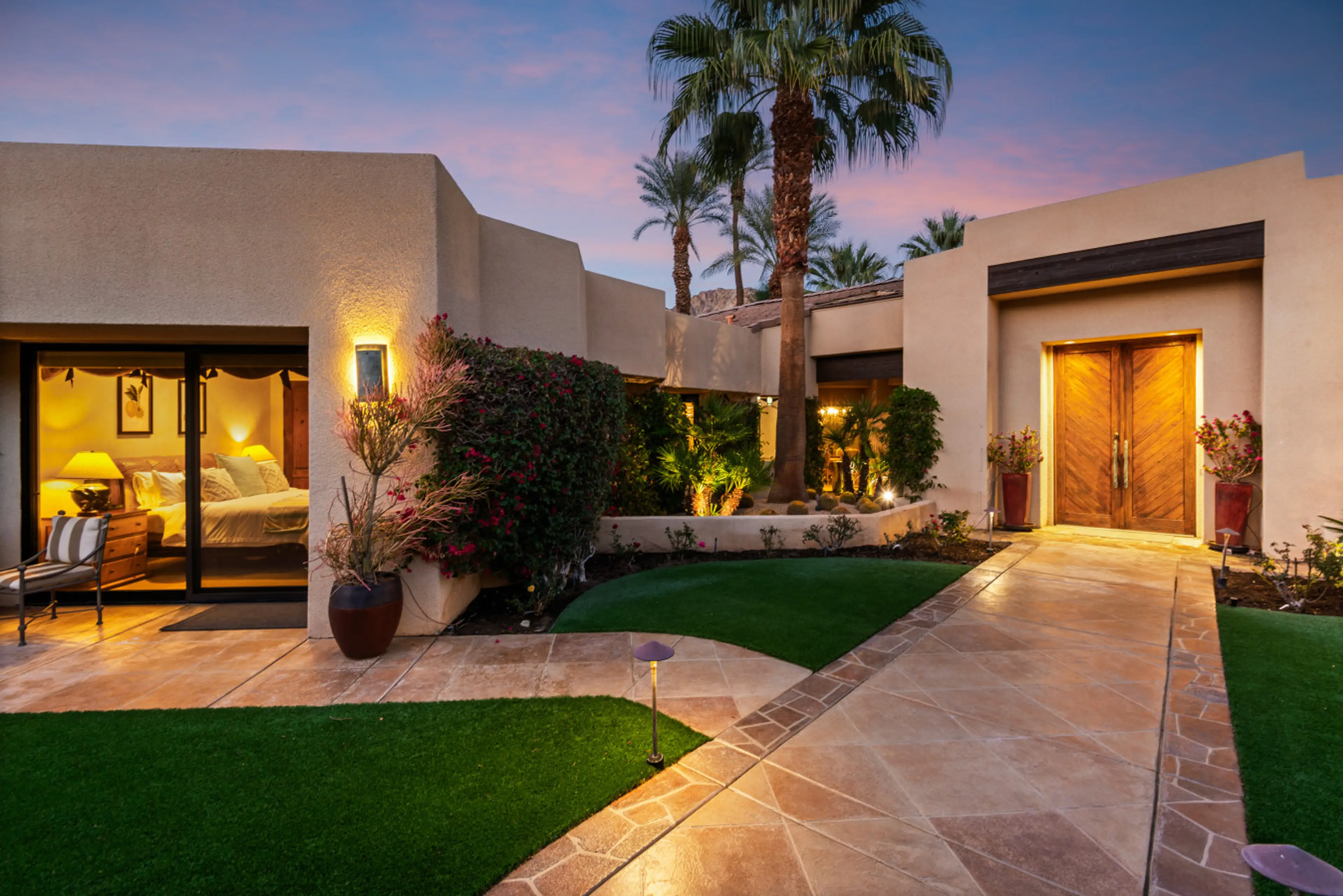 A beautifully illuminated private entrance framed by lush landscaping and palm trees, creating a warm, inviting arrival with a perfect blend of desert elegance and resort-style charm at dusk.