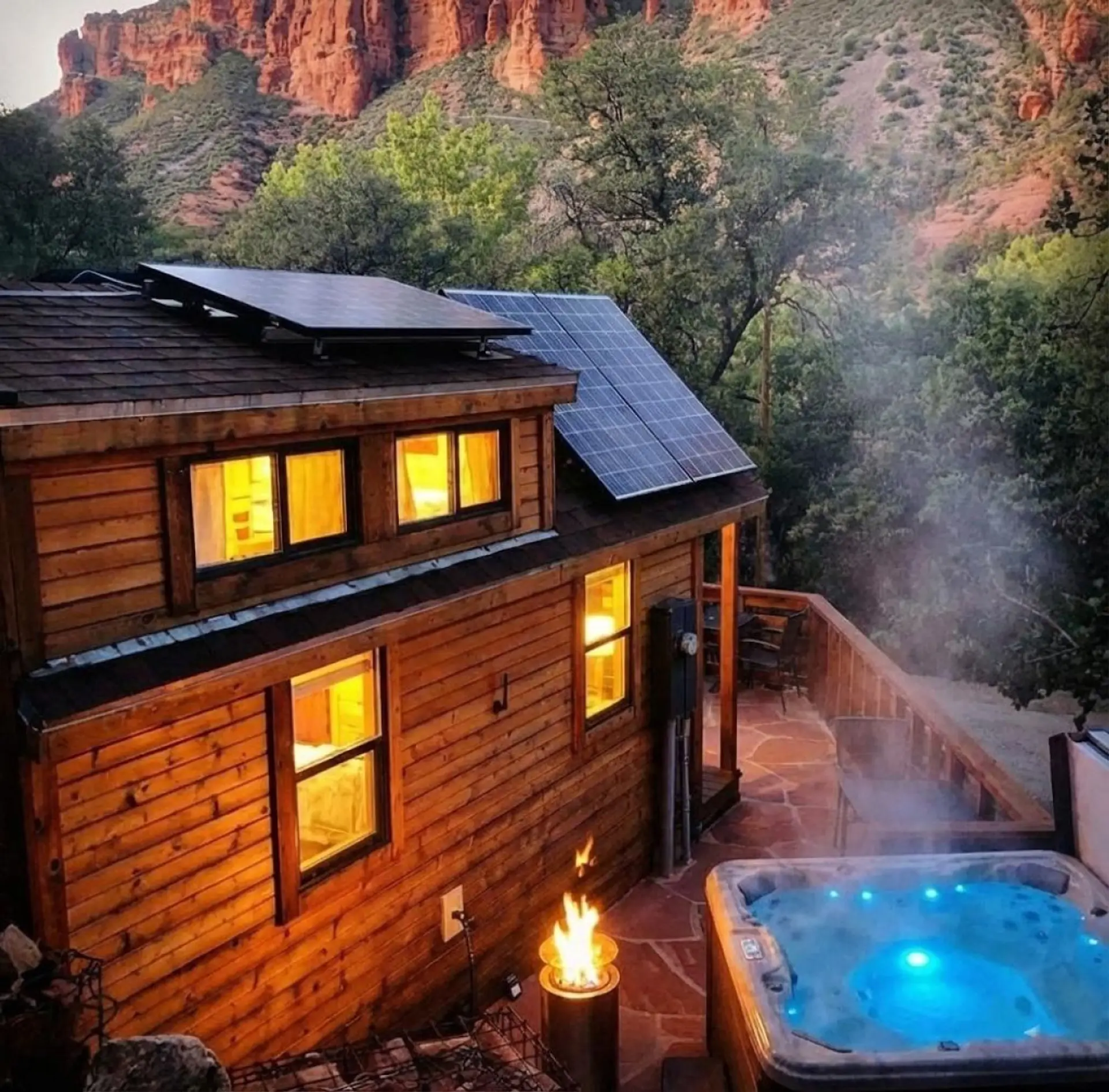 Steam rises off the hot tub as string-lit windows glow against the red rock cliffs above — solar panels on the roof mean this magical scene runs on clean energy, so you can soak guilt-free under the stars.