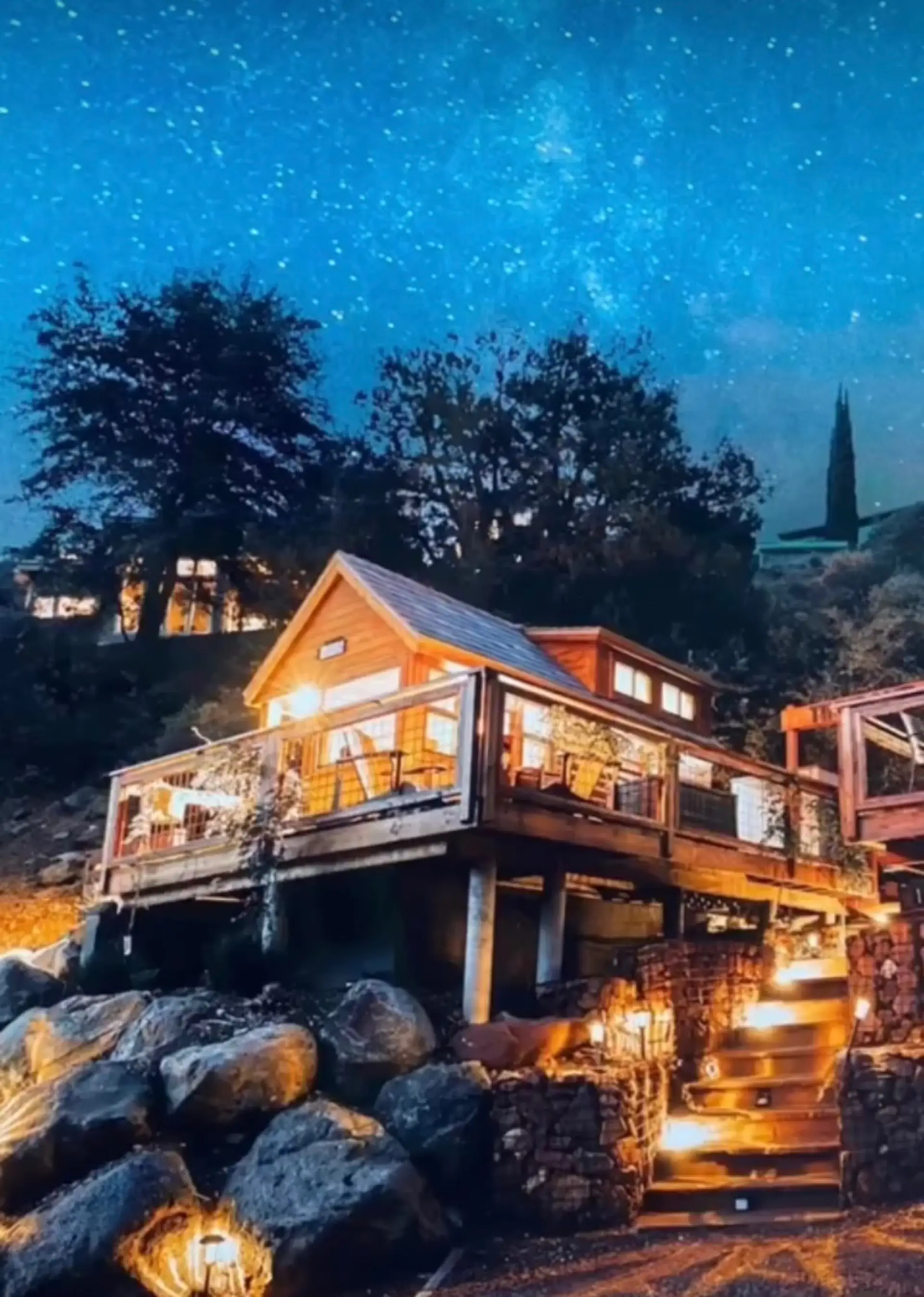 Perched above a rocky hillside and lit from within, this cabin glows warmly beneath a sky full of stars — a lit stone staircase winds up through the boulders to greet you, making the walk home each night part of the magic.