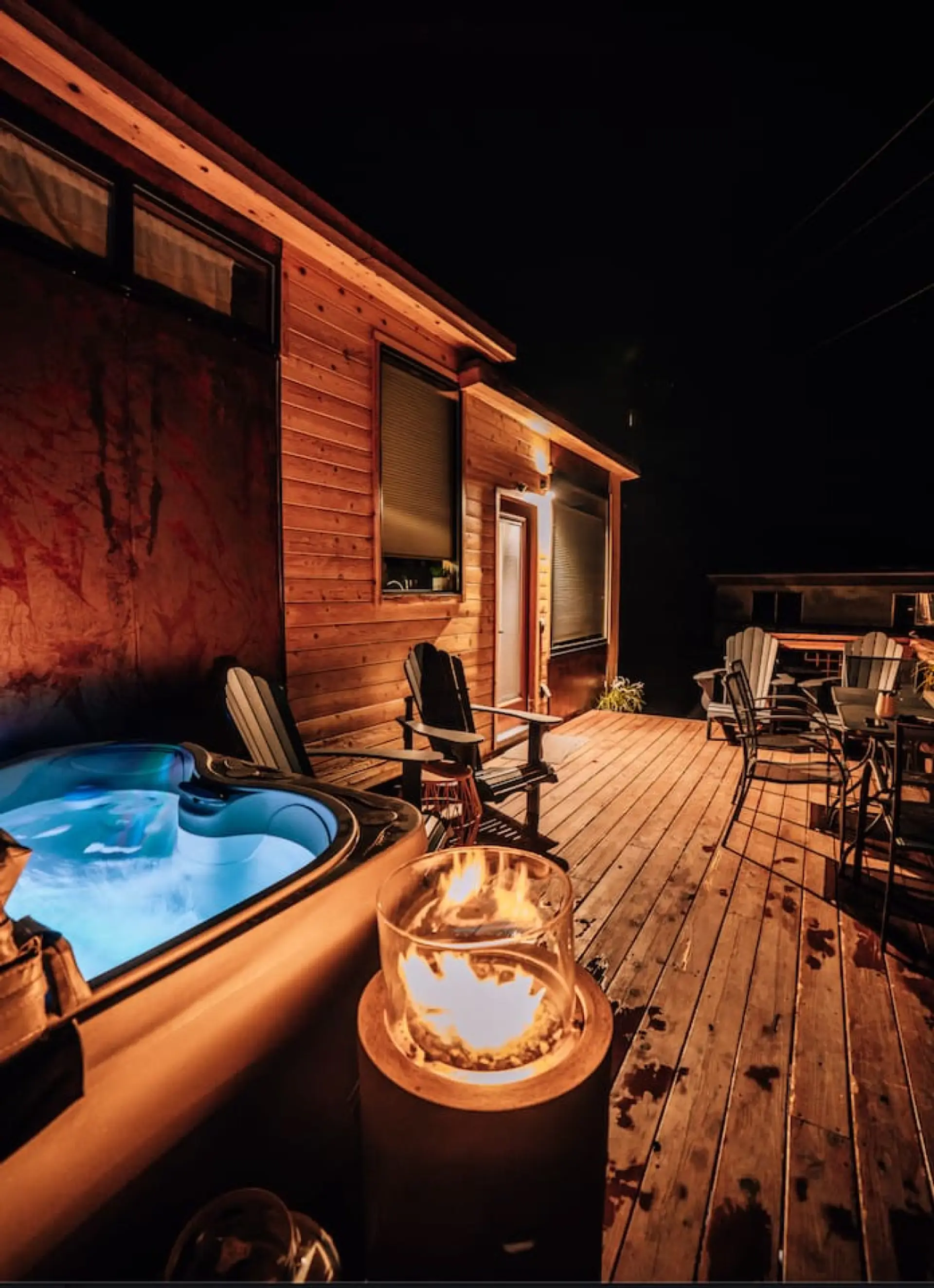 Private hot tub glowing at night beside a rustic metal art wall, fire pit nearby.