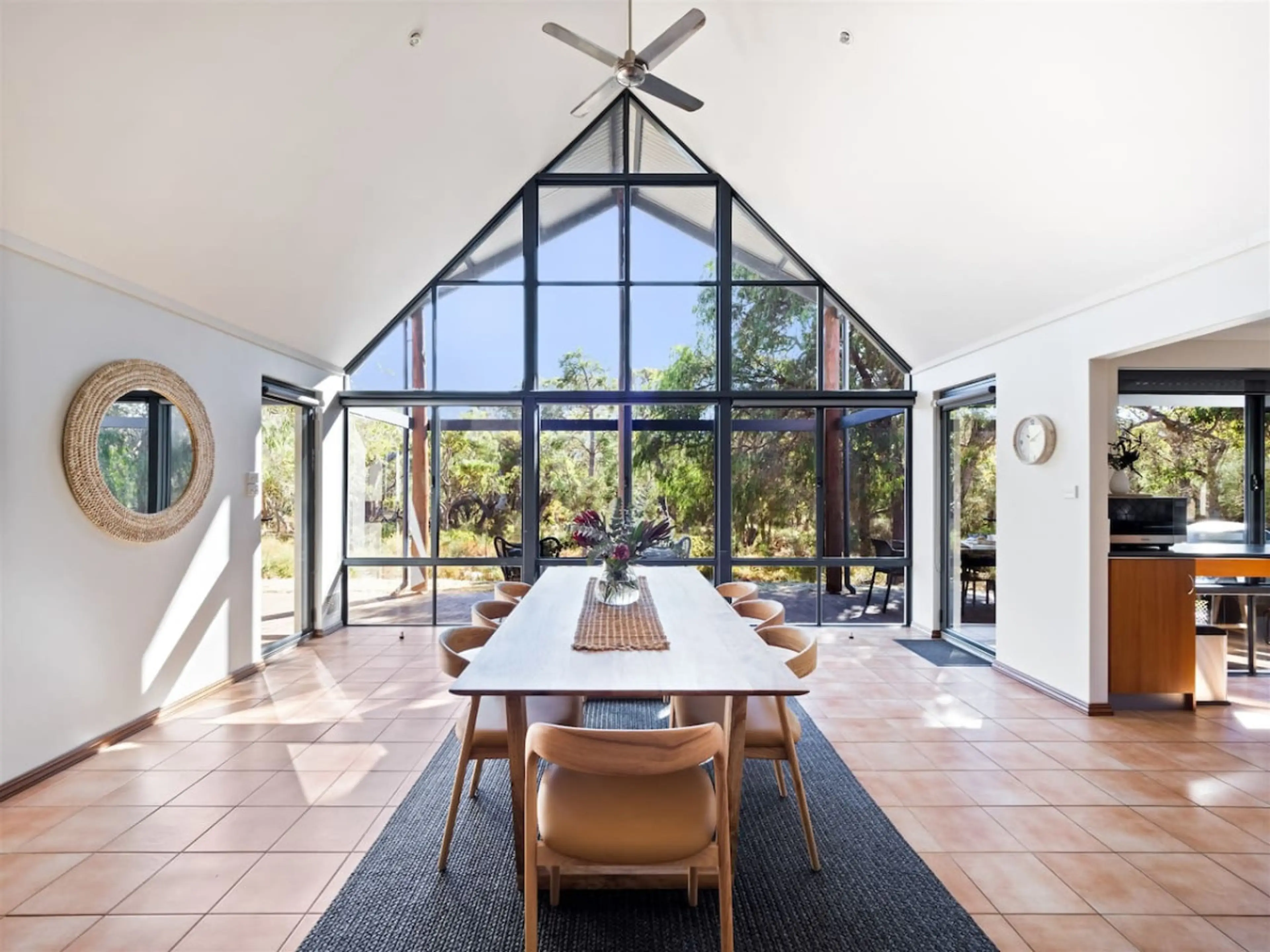 A high vaulted ceiling Dining Room with large windows. The dining table is made of light-colored wood and is surrounded by brown leather chairs. A black and white patterned rug sits under the table, with a round mirror hangs on the wall.