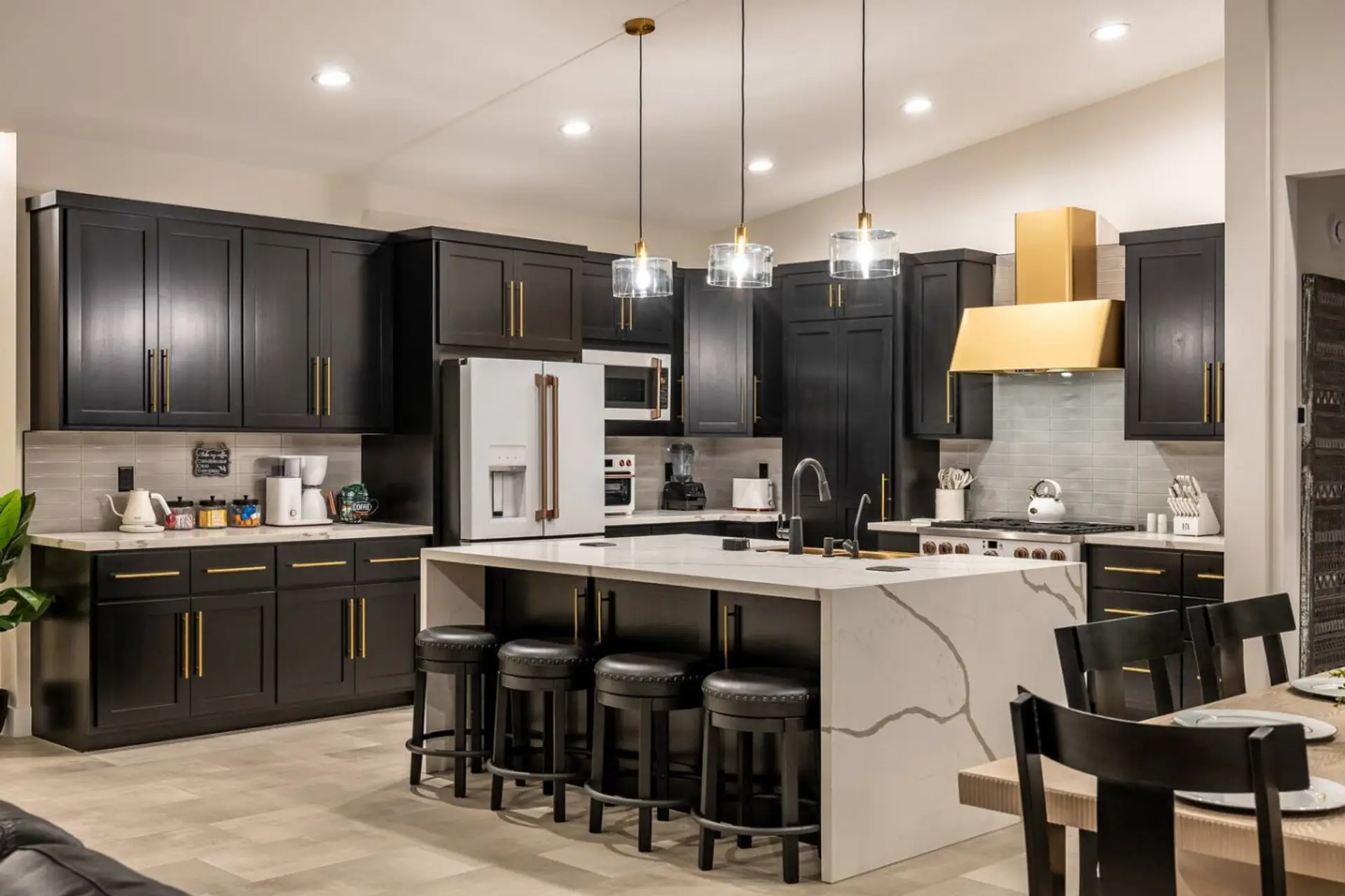 Fully stocked chef's kitchen — dark shaker cabinets, marble-veined waterfall island, gold range hood, and premium appliances. Everything ready for a family feast.