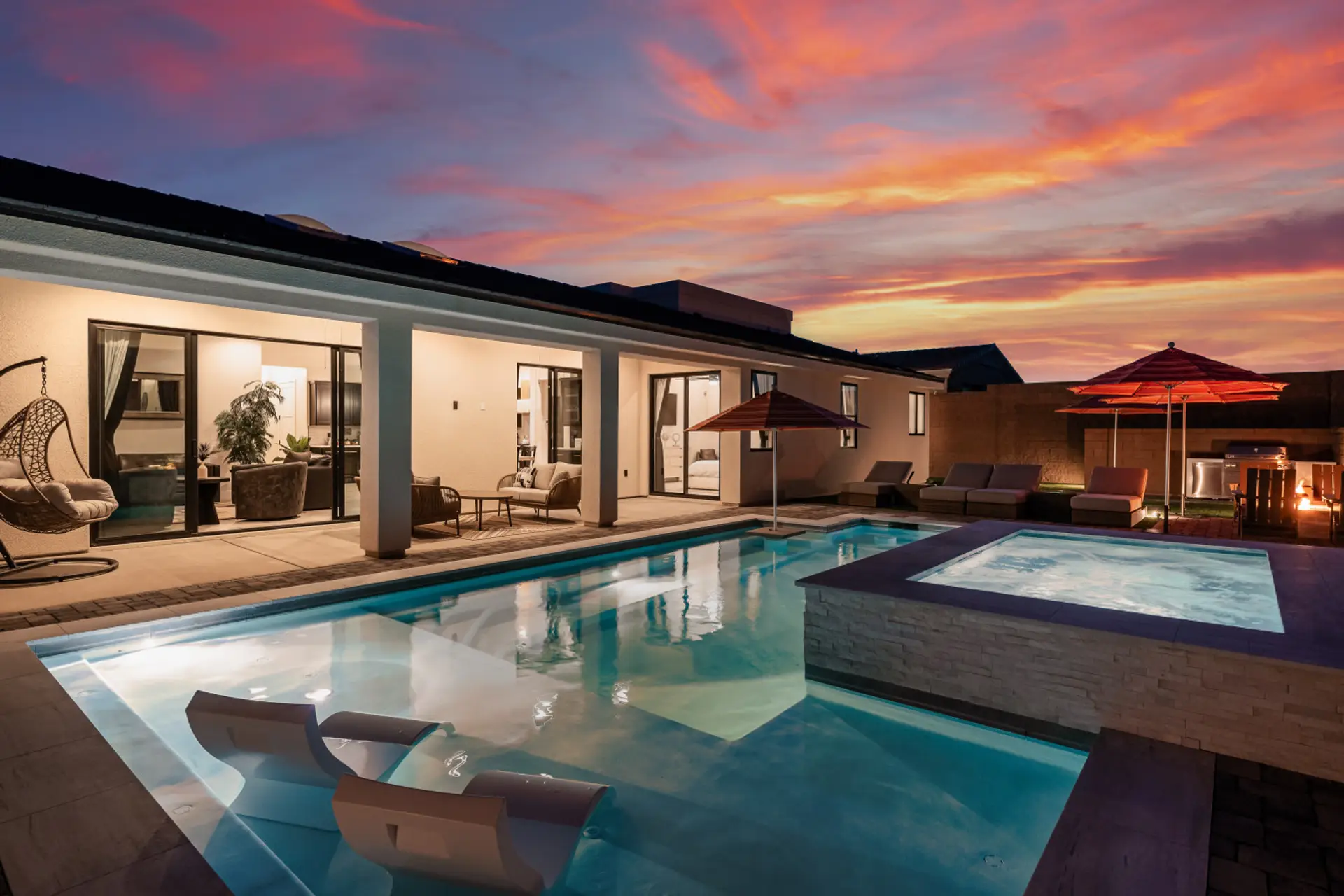 Twilight backyard — lit pool with in-water loungers, raised spa, fire pit glowing at dusk, and hanging egg chair. The shot guests screenshot before they book.