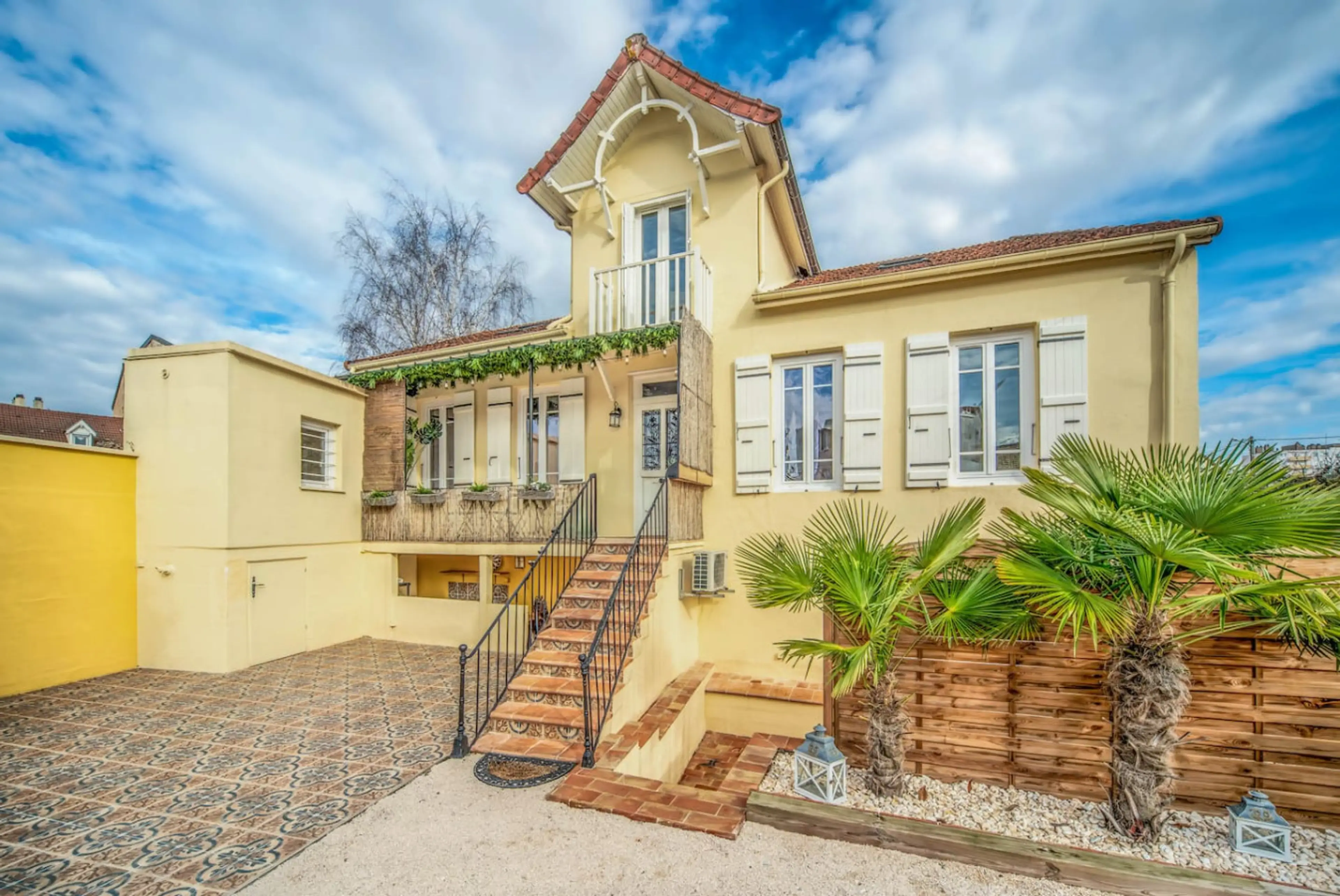 Maison de charme jaune pâle avec terrasse pavée, palmiers méditerranéens et accès par escalier en bois