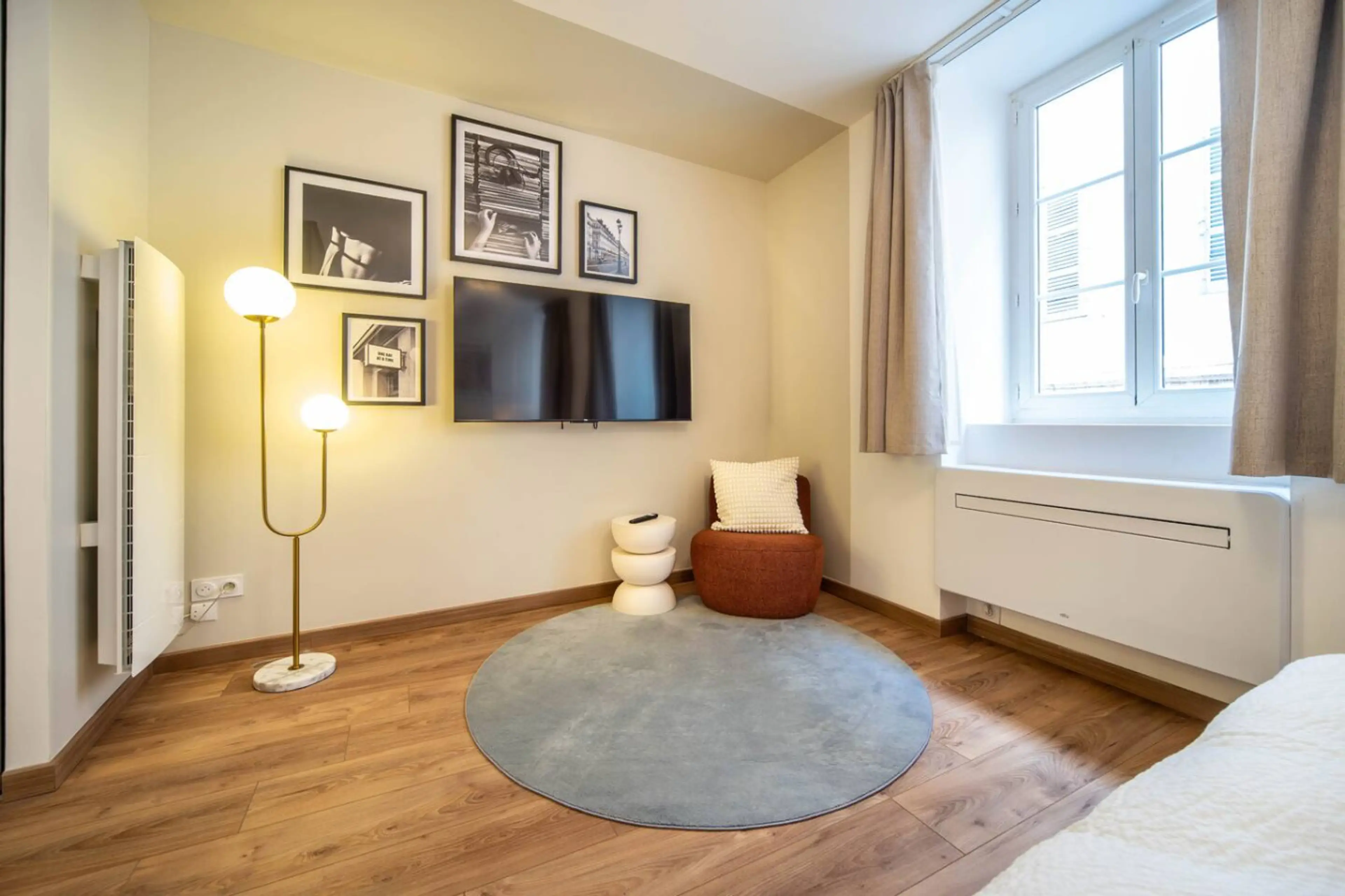 Salon cosy avec télévision murale, tapis gris, lampadaire design et grandes fenêtres lumineuses