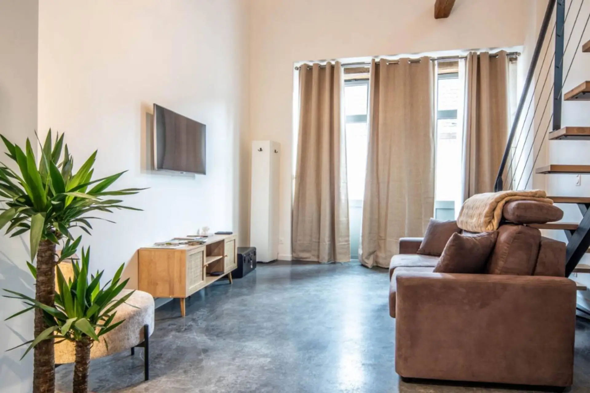 Spacieux séjour lumineux avec canapé confortable, plantes vertes, escalier et grandes fenêtres donnant sur la ville