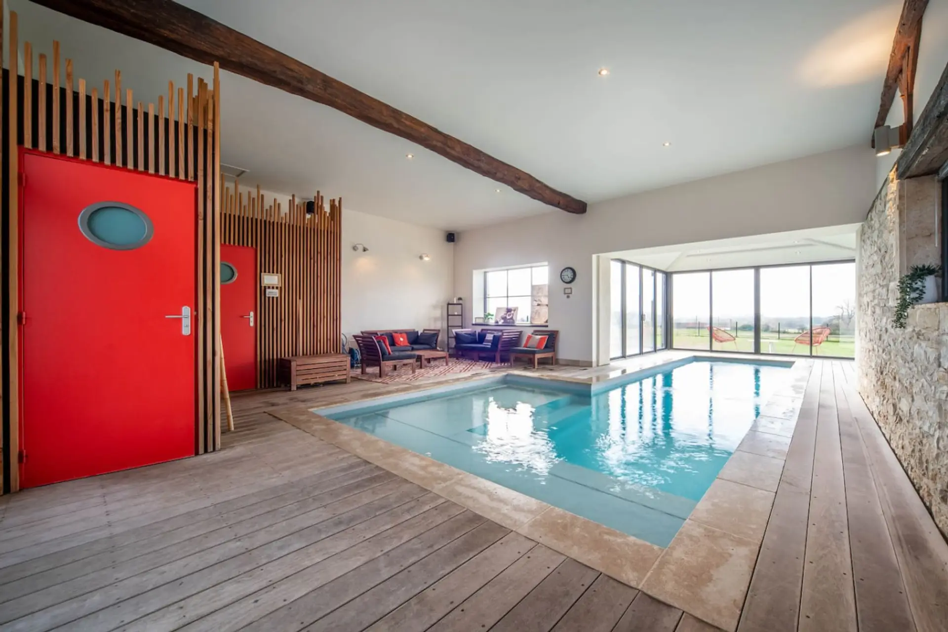Espace piscine intérieure spacieux avec bassin bleu, terrasse en bois, baies vitrées et salon confortable