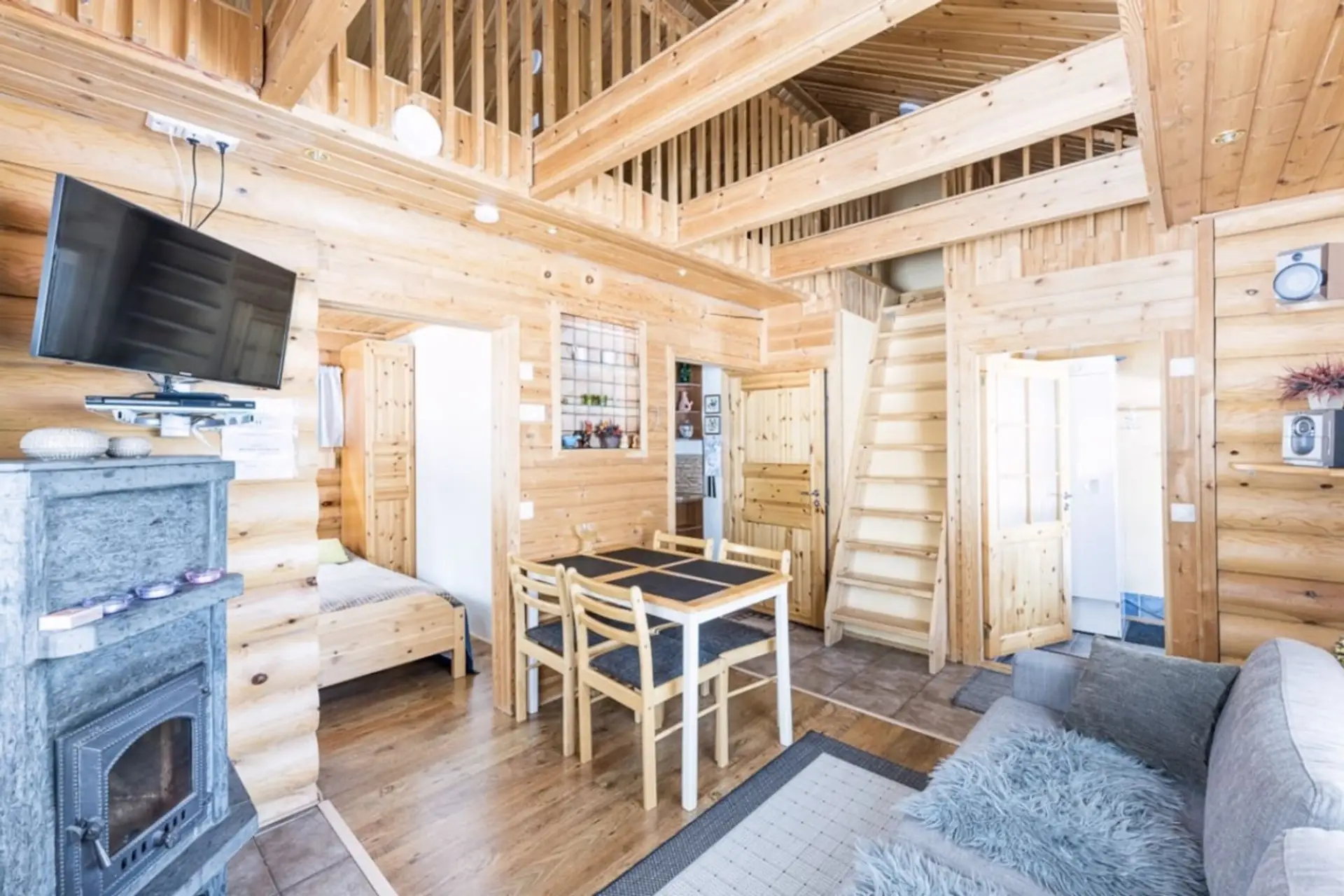 Bright open-plan living space with a stone fireplace, wall-mounted TV, dining table and stairs up to the loft.