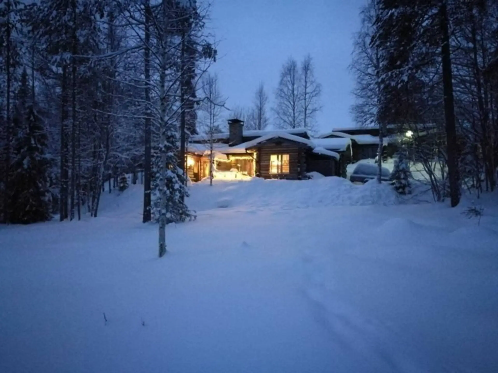 The log cabin lit up on a winter evening, surrounded by deep snow and pine trees.