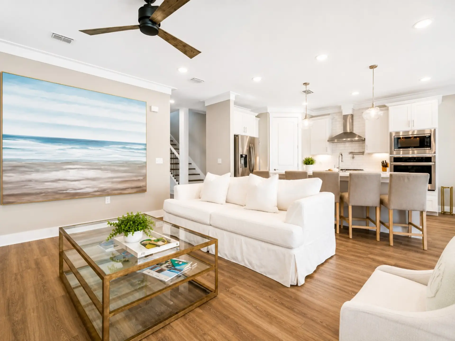 Living Room – Open-concept great room with a coastal ocean mural, plush white sofa, and gold-framed glass coffee table flowing into the kitchen.