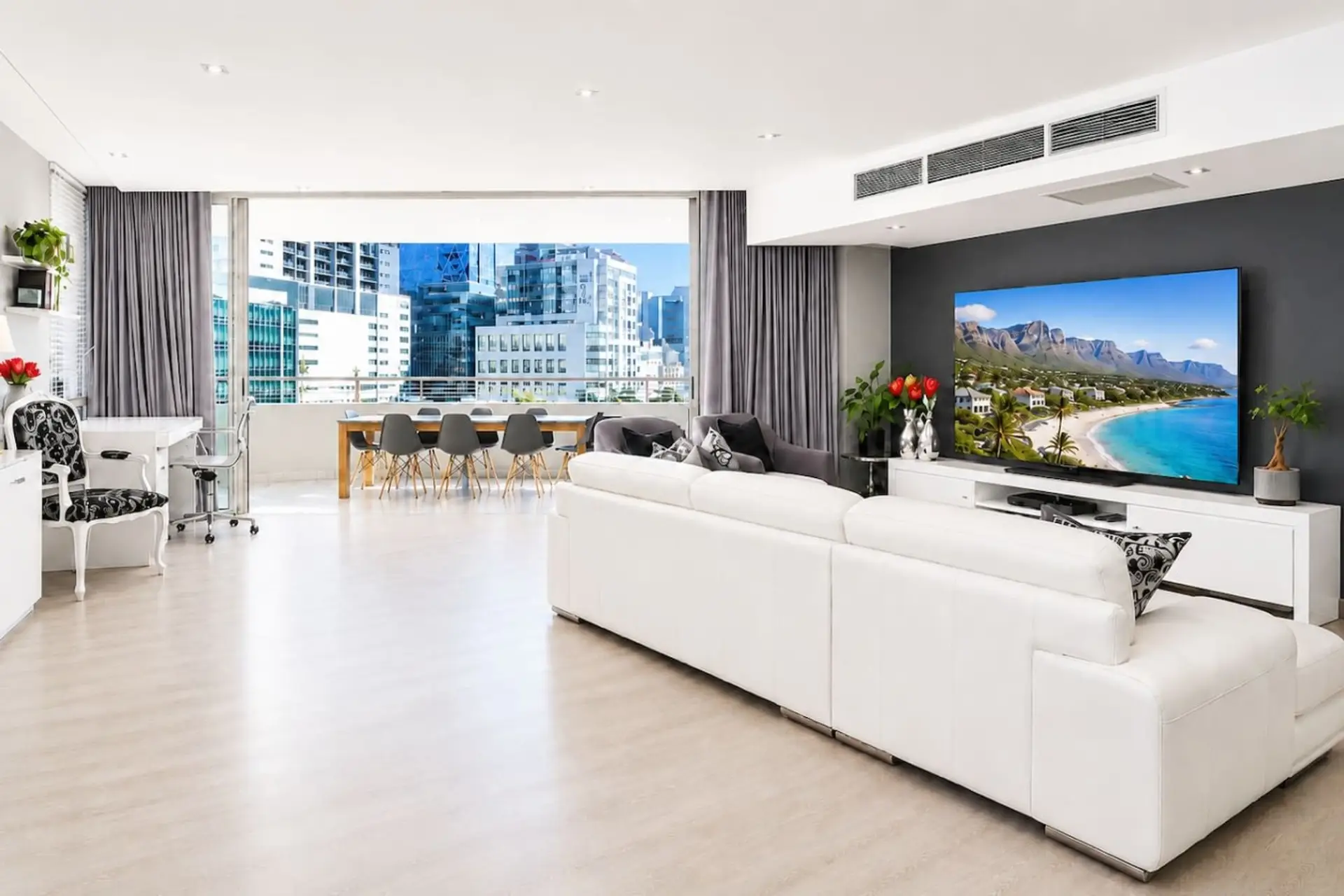 Open-Plan Living Area: Bright and spacious open-plan living area designed for comfort and entertaining, featuring stylish furnishings, a large smart TV, dedicated dining space, and expansive city views through oversized windows.