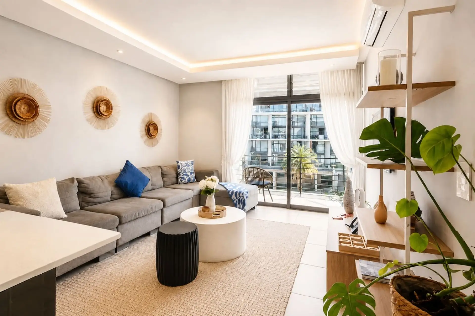 Living Room: Airy lounge featuring a generous corner sofa, textured rug, sculptural coffee tables, woven wall pieces, and curated shelving, enriched with leafy greenery and wide glass doors that bring in abundant daylight.