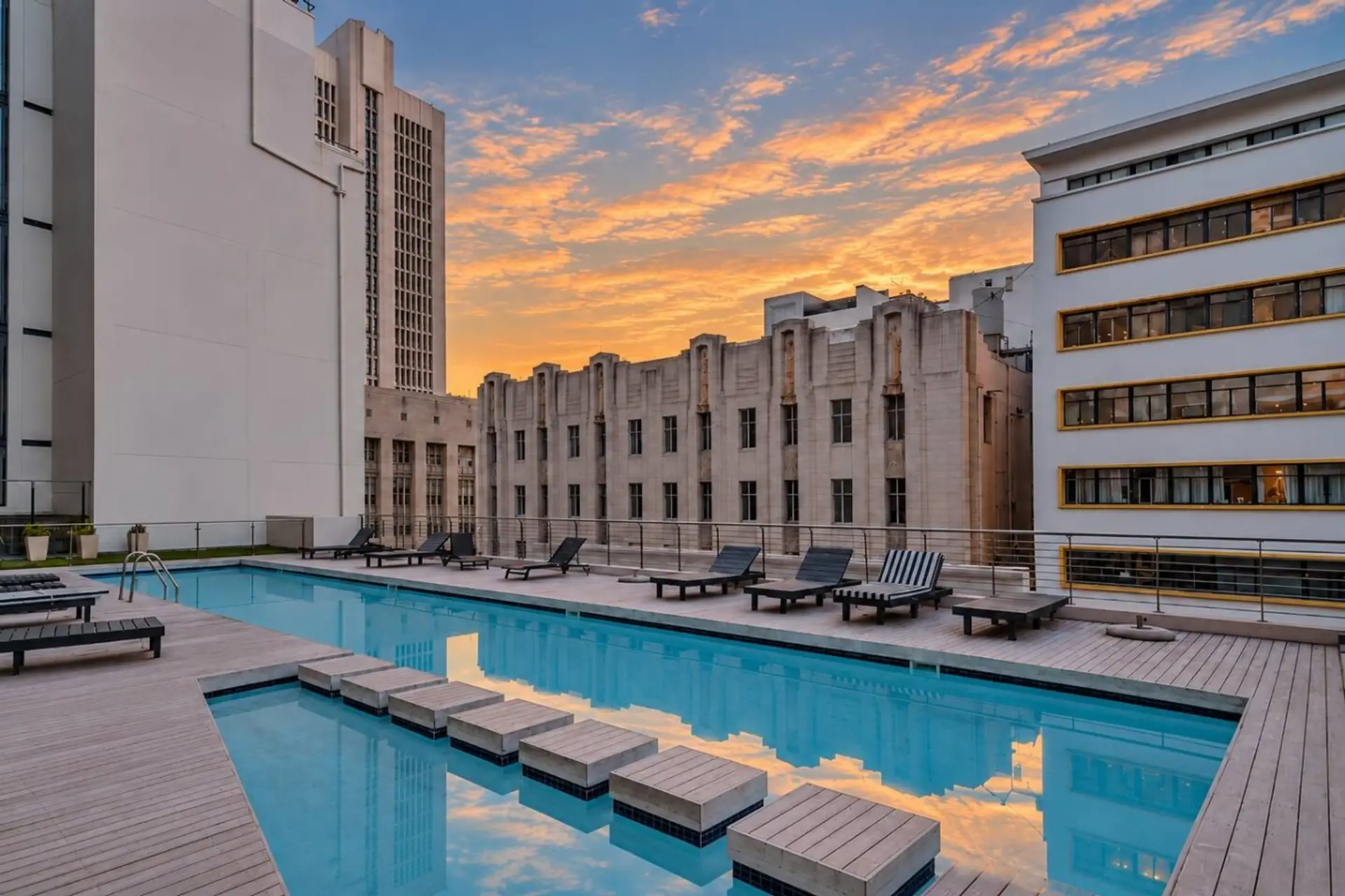 Pool Deck: A distinctive split-level pool design features stepping platforms across the water, surrounded by reclining sunbeds and spacious decking. Historic façades and modern buildings create an interesting architectural backdrop at dusk.