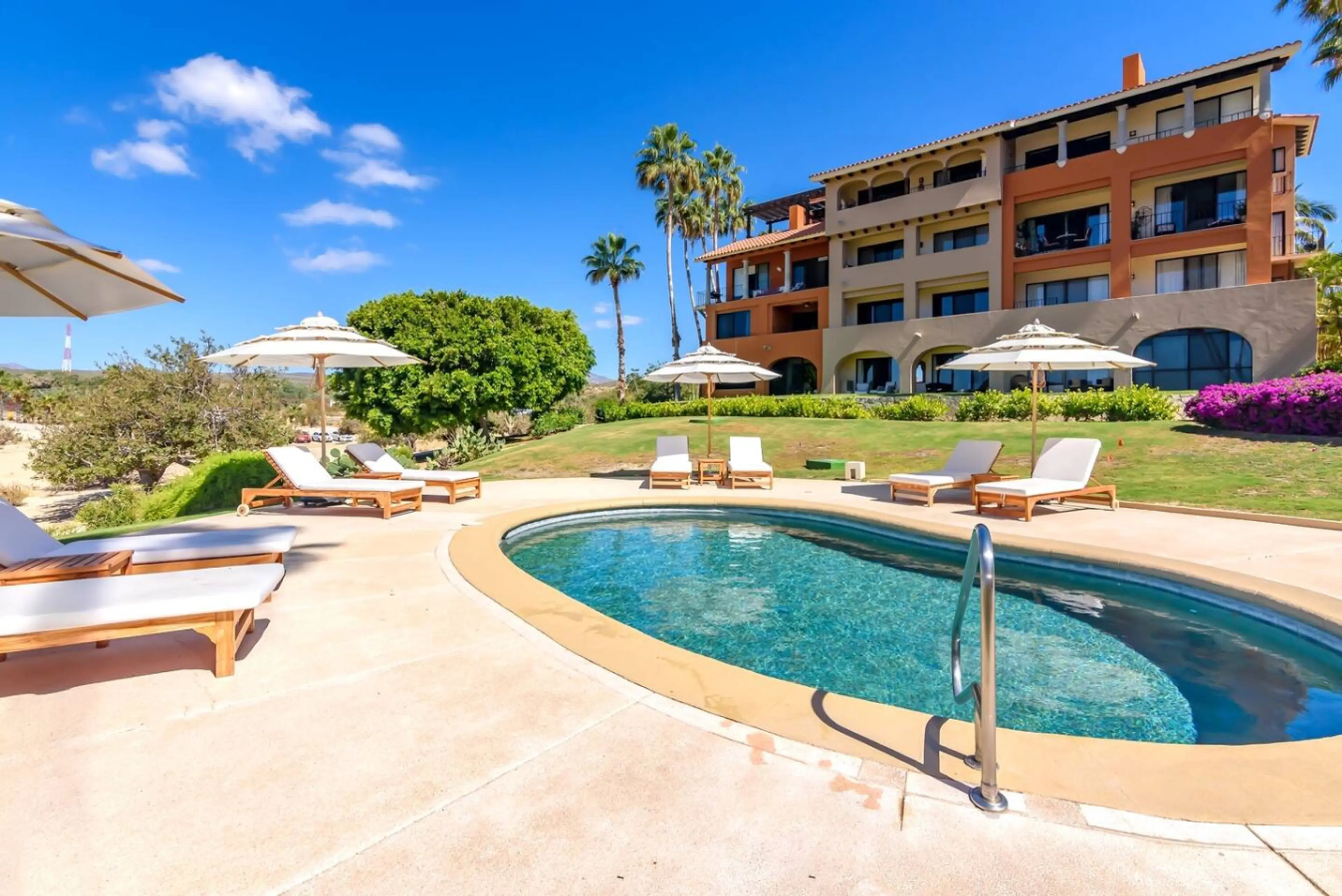 A modern apartment complex with a shared pool, sun loungers, and palm-lined gardens.