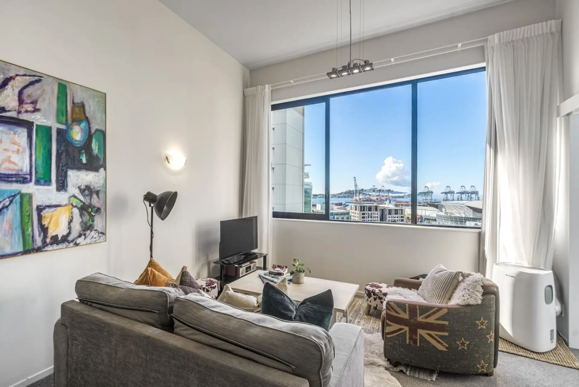 The living area boasts high ceilings, sheer & blackout curtains, air conditioning, and opening windows. The lounge offers a comfortable sofa, occasional chair, bean bag, television, and stunning views out to Rangitoto Island.