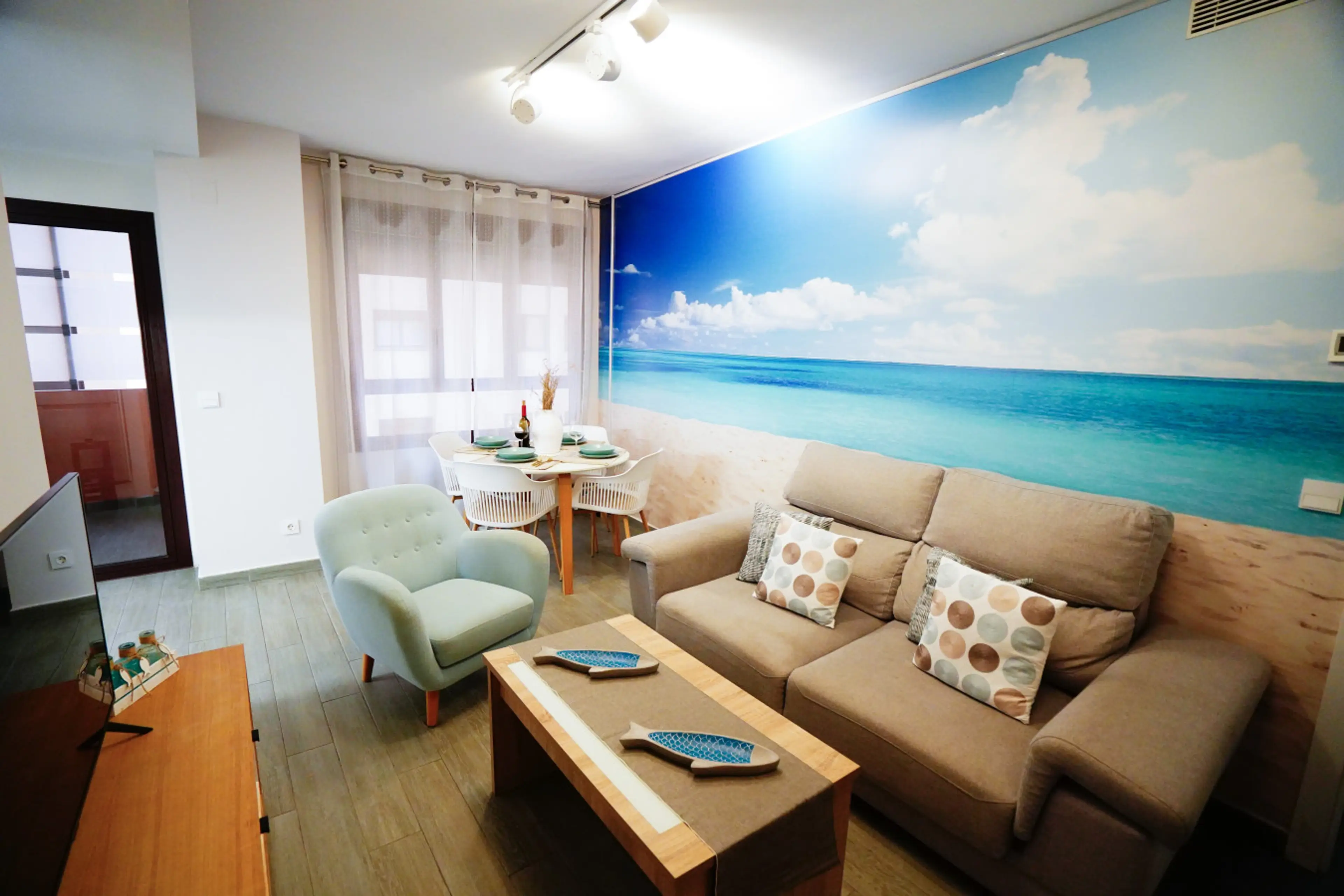 Vista general del salón-comedor con sofá en tonos neutros, sillas azules y mural panorámico del mar. El espacio destaca por su luz natural y estilo moderno con toques costeros. Ideal para reuniones o momentos de descanso en un entorno luminoso.