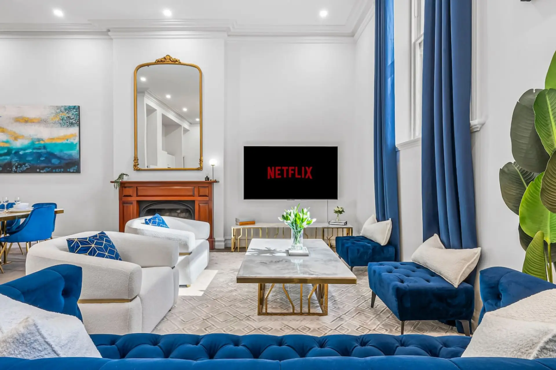 Elegant living room with plush blue and white seating, marble coffee table, fireplace with gold mirror, and a smart TV with Netflix. Bright natural light, tall curtains, and refined decor create a cozy yet luxurious atmosphere.