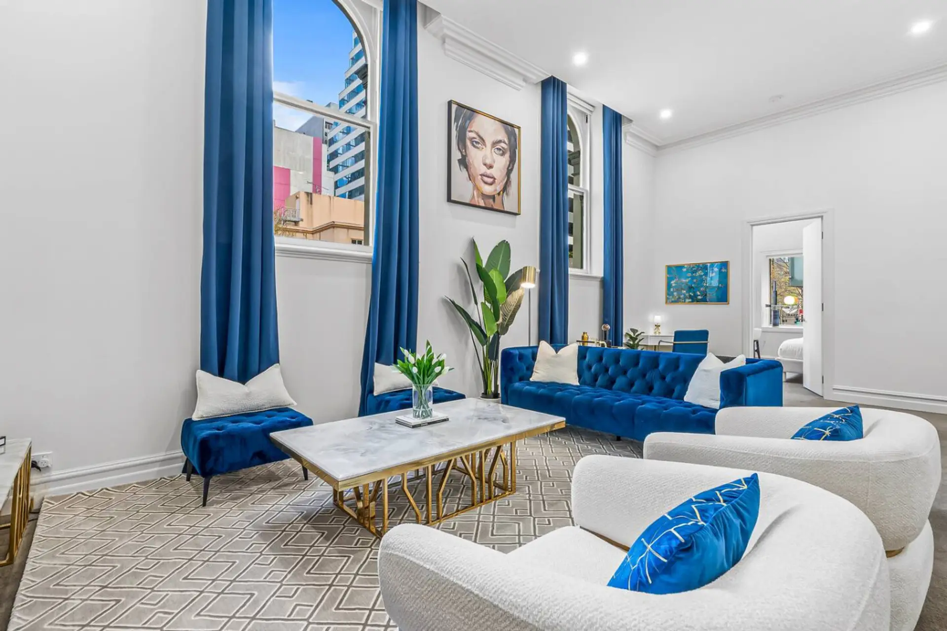 Bright and elegant living room with plush blue velvet sofas, marble coffee table, tall arched windows, and city views. A stylish space designed for comfort, natural light, and relaxed conversations.