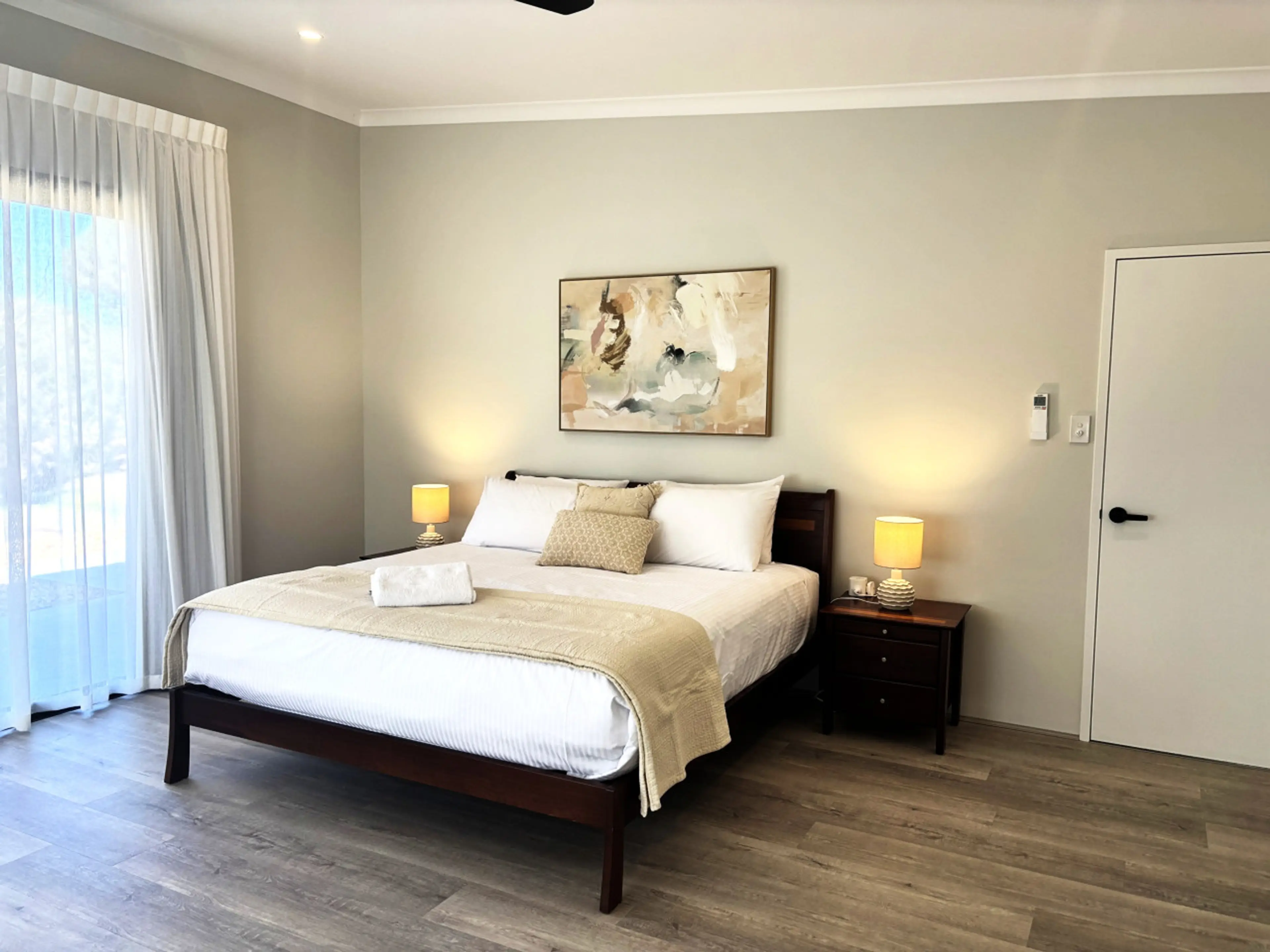 Spacious king master suite with warm timber tones, soft neutral styling and gentle bedside lighting. Wake to filtered morning light through sheer curtains, stretch out on a premium king bed with crisp white linen, and enjoy the calm, uncluttered space des