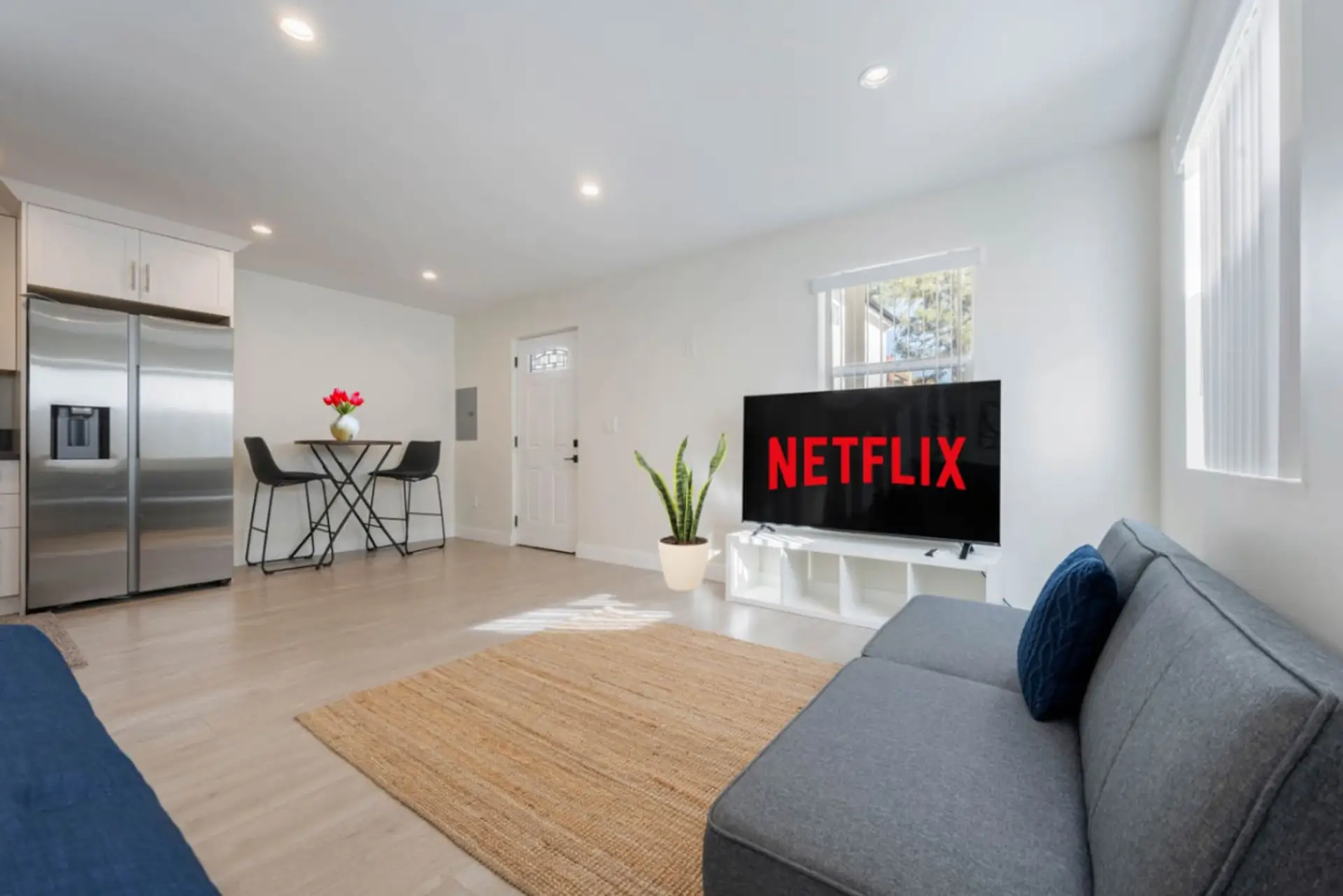 A cozy living area with a gray sofa bed, large TV ready for Netflix, a small dining nook, and a stainless steel fridge.