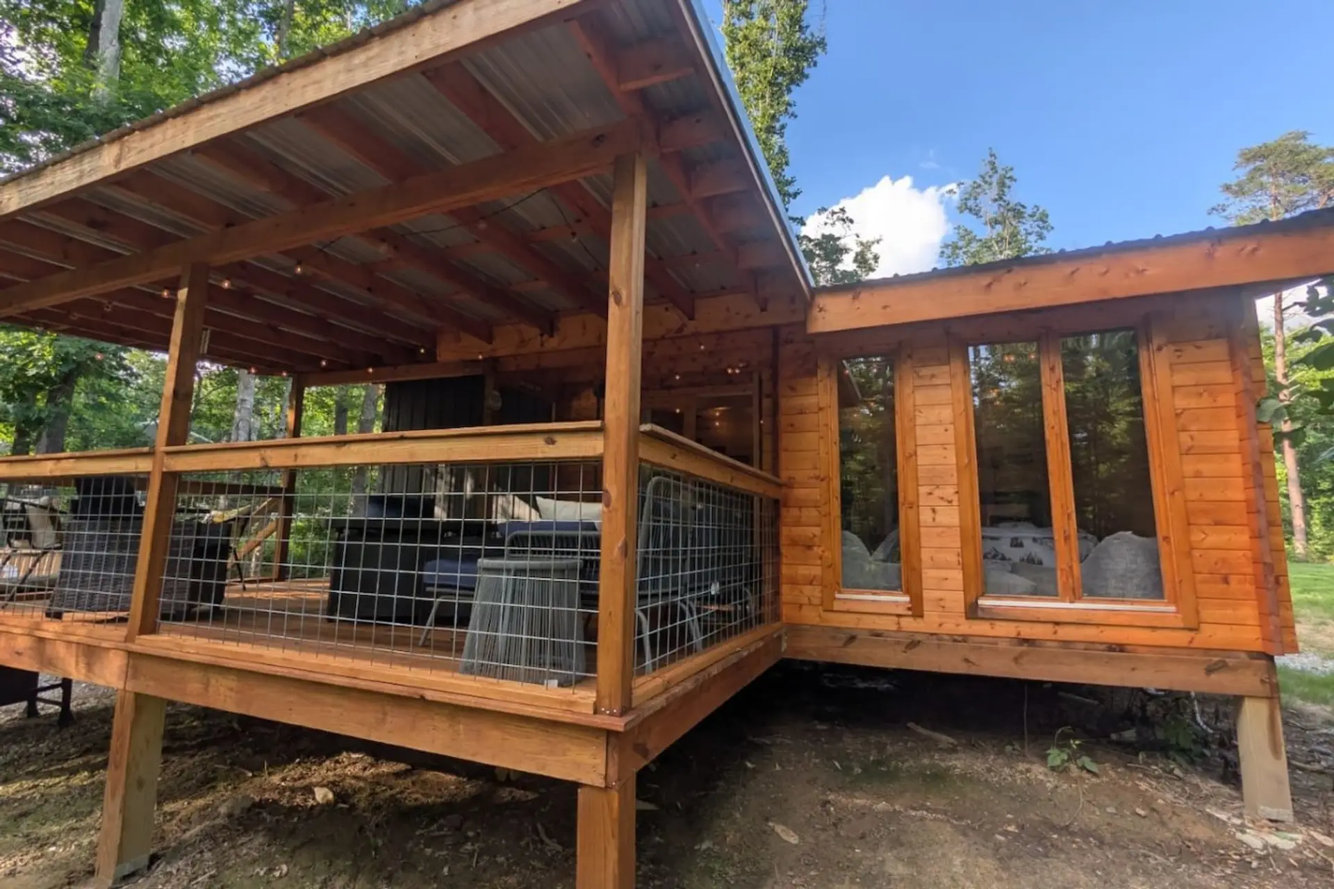 The Nordic Spruce Cabin in daylight, showing the large covered porch and the floor-to-ceiling bedroom windows that look right out into the woods.