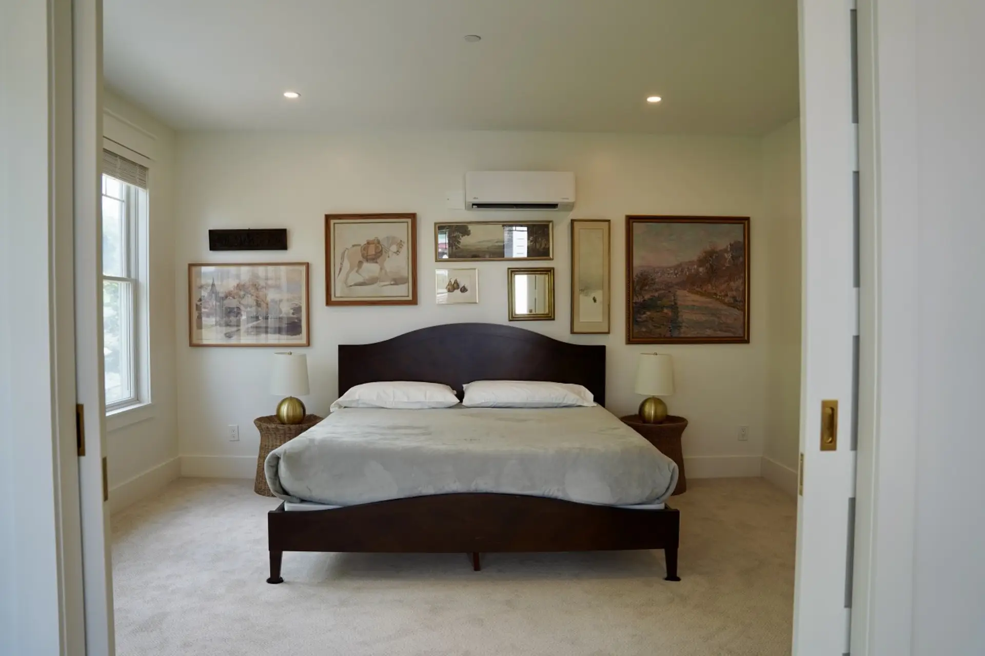 You now enter a serene bedroom centered around a rich wood king bed. You will experience a curated gallery of vintage art, matching brass lamps on woven nightstands, and soft carpet underfoot, all bathed in natural light from the window.