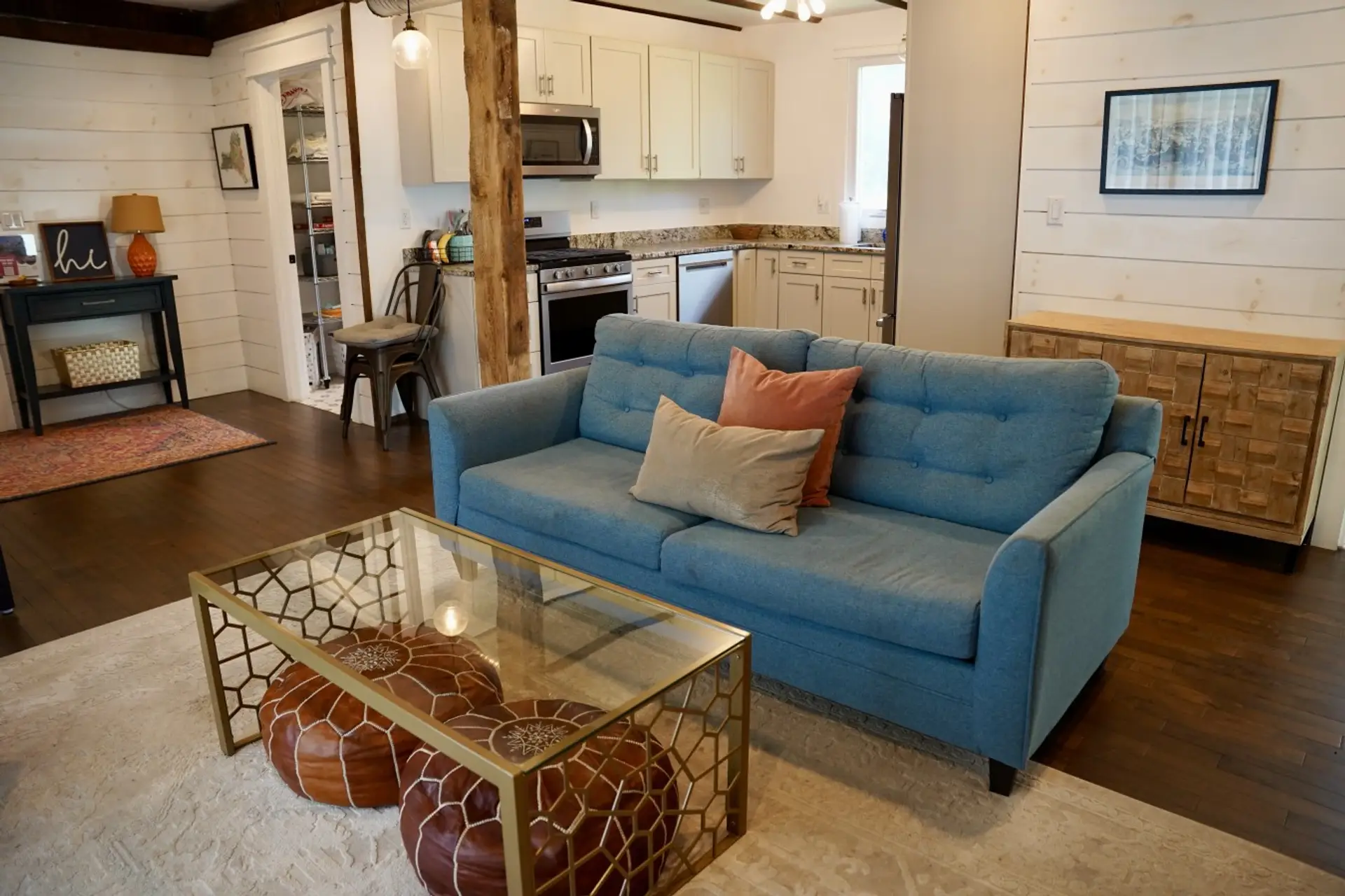 Open concept living space with a comfy blue sofa, leather Moroccan poufs, and original barn beams overhead. The fully equipped kitchen sits just steps away, making this bright, character filled space perfect for relaxing or entertaining.