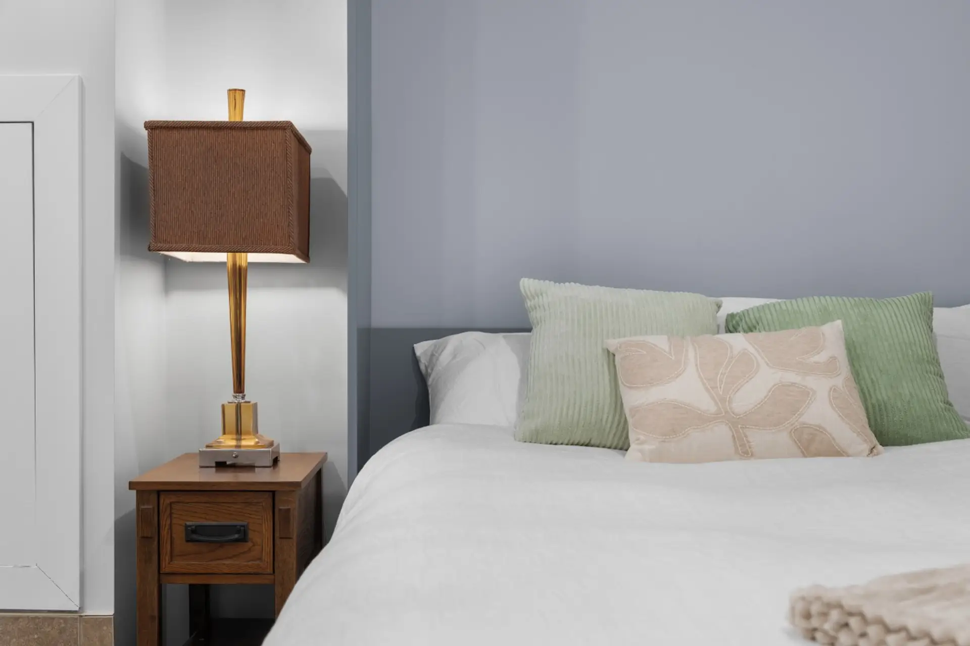 Cozy private lower-level bedroom in a modern Calgary townhouse. Features a comfortable Queen bed, stylish textured sage and patterned accent pillows, and ambient nightstand lighting.