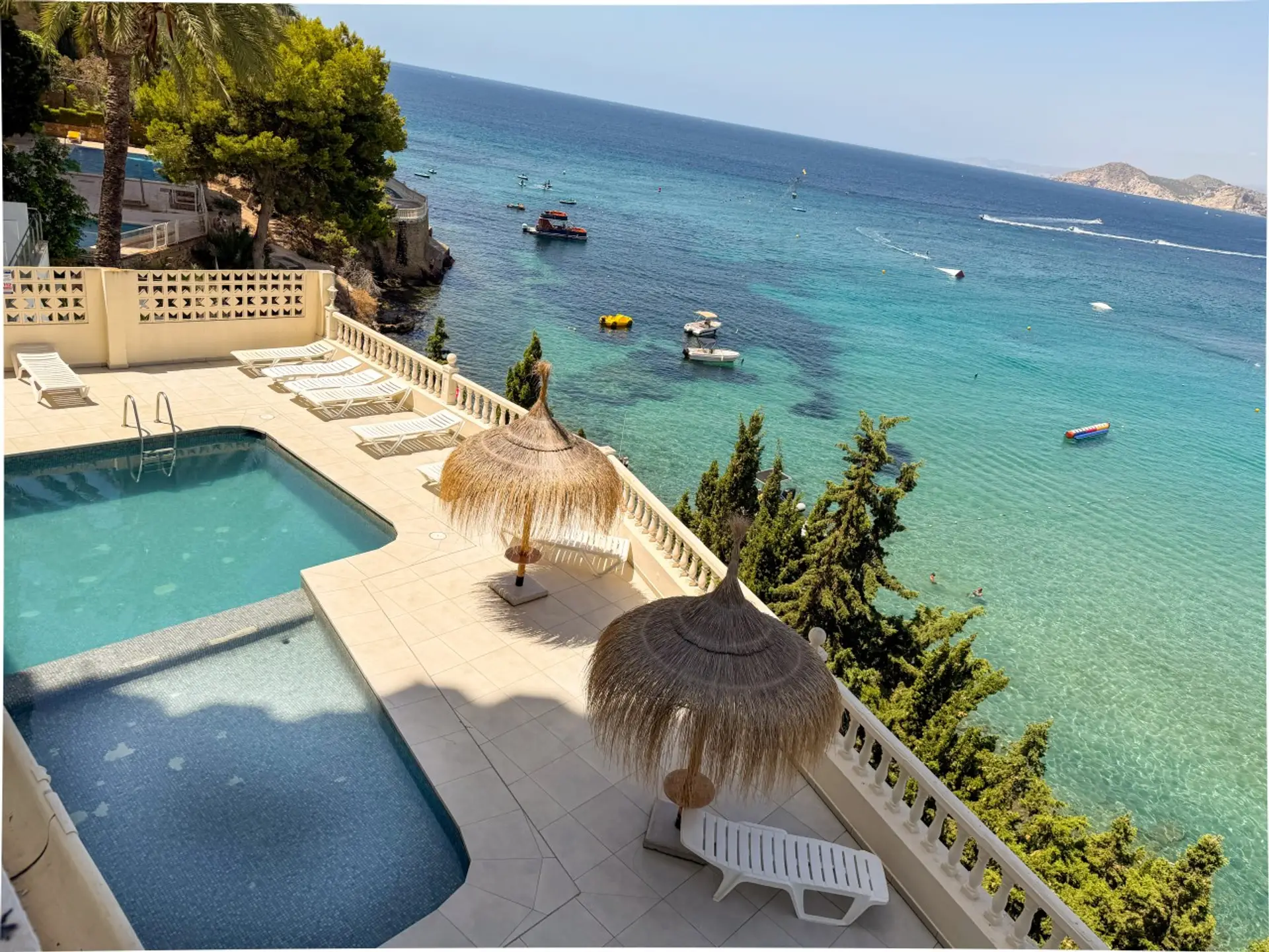 Vista elevada de la piscina comunitaria exterior con sombrillas de paja y el mar de fondo.