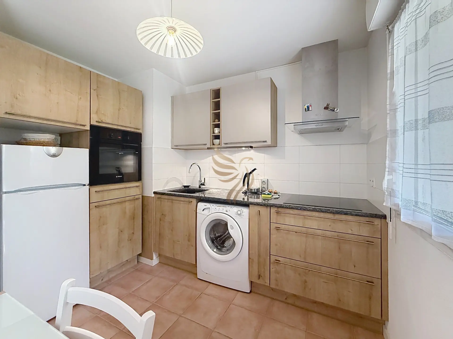 Matin tranquille dans cette cuisine chaleureuse : la lumière filtre par le rideau pendant que vous préparez un café sur le plan sombre, tout est pratique et rangé, du four encastré au lave-linge discret.