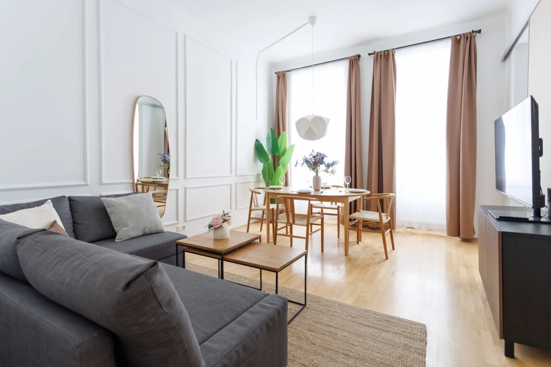 Helles Wohnzimmer mit grauem Ecksofa, Esstisch mit vier Stühlen, Flachbild-TV, Parkettboden und hohen Fenstern in der Wohnung in Wien-Ottakring (1160)