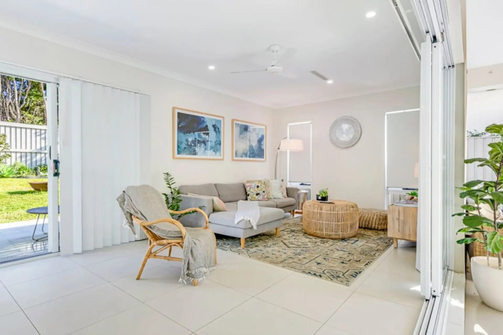 Sunlit living room with sectional sofa, rattan armchair, ceiling fan and sliding doors to the backyard — coastal calm from the moment you walk in.