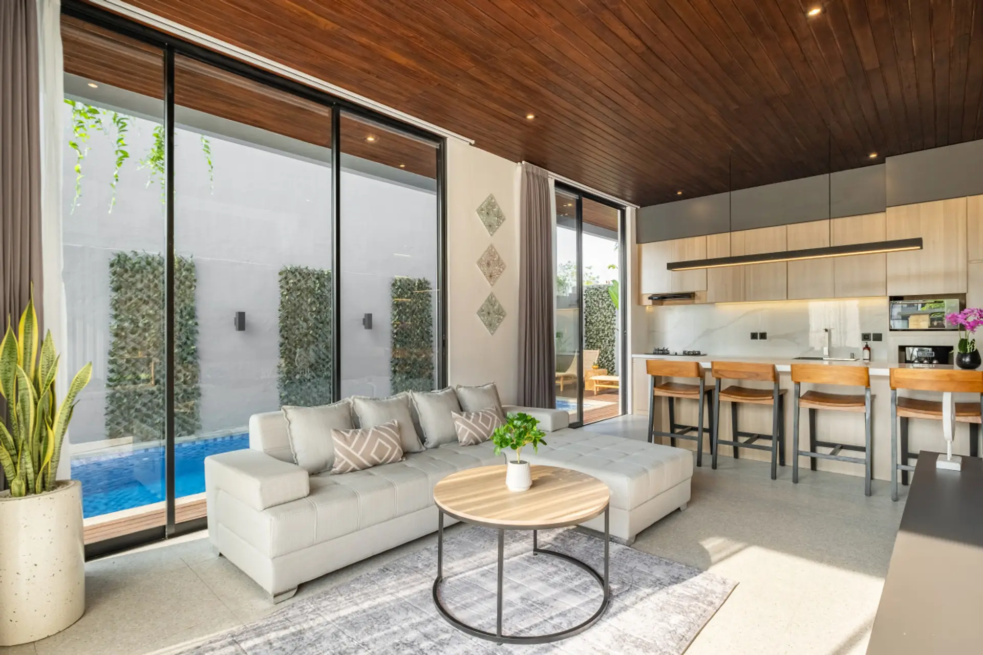 A bright, modern open-plan living area with a cozy sofa, stylish kitchen with bar seating, and large glass doors overlooking a private pool.
