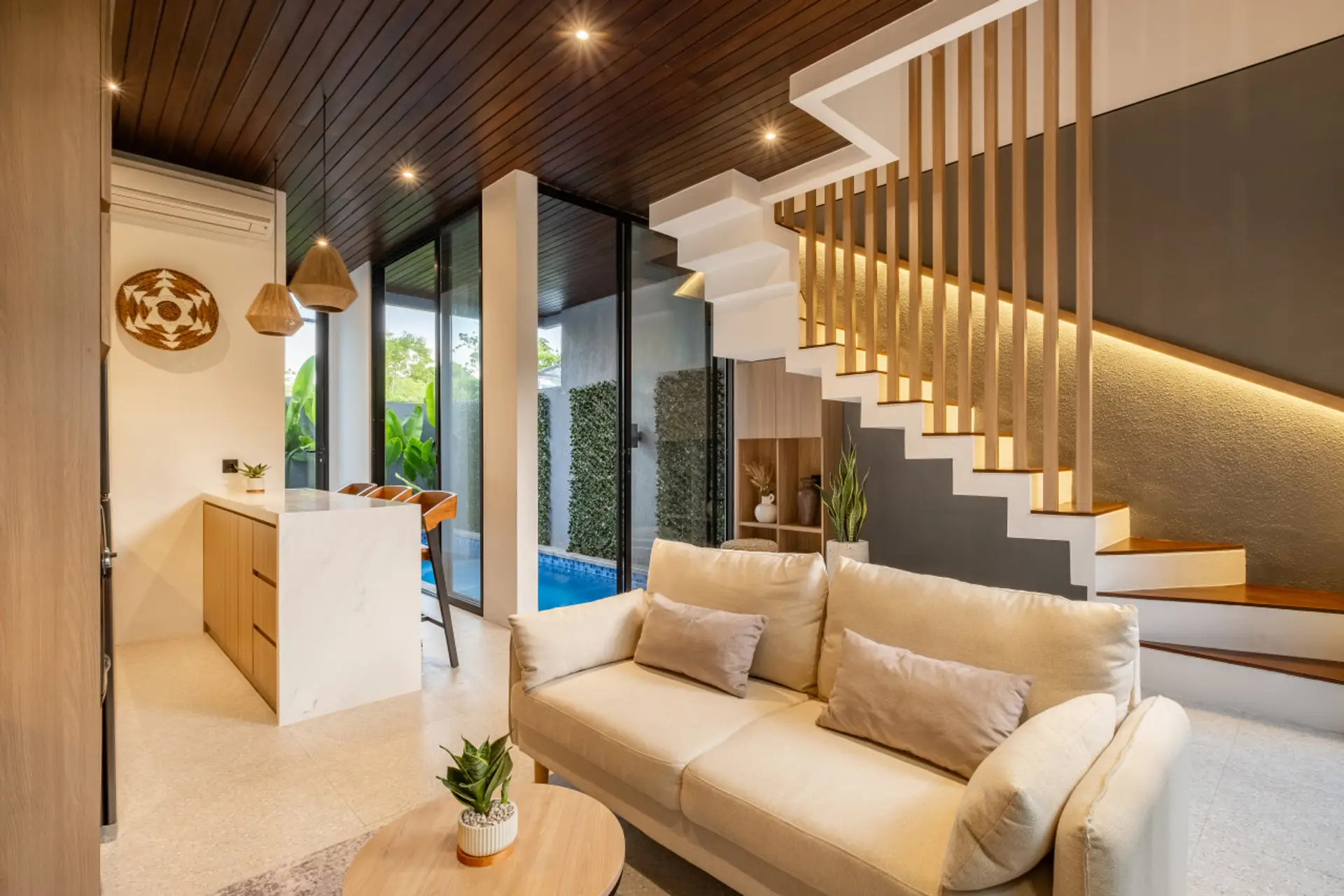 A bright, modern open-plan living area with a cozy sofa, stylish kitchen with bar seating, and large glass doors overlooking a private pool.