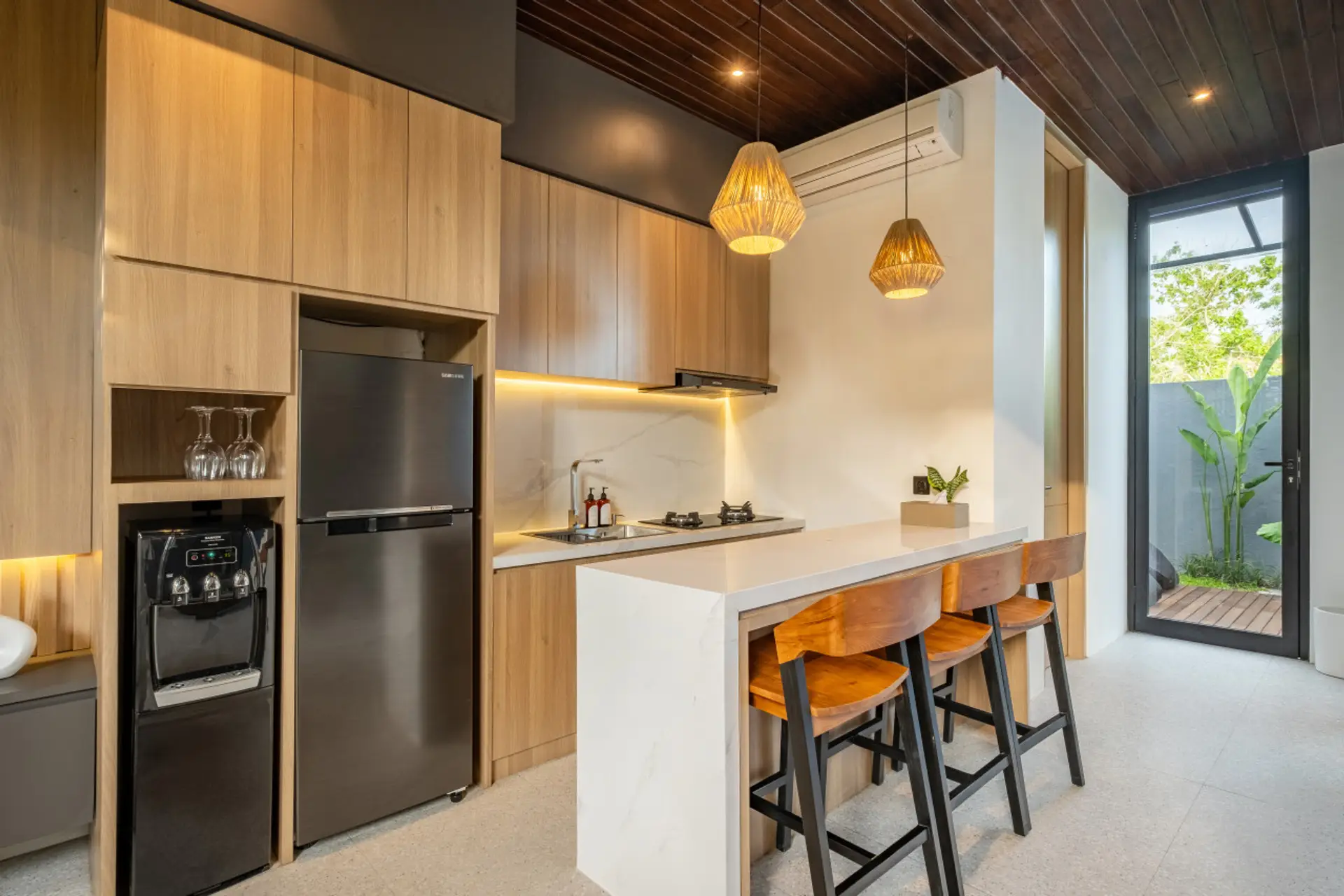 A modern kitchen with light wood cabinetry, a sleek island with bar stools, built-in appliances, and large glass doors providing natural light.