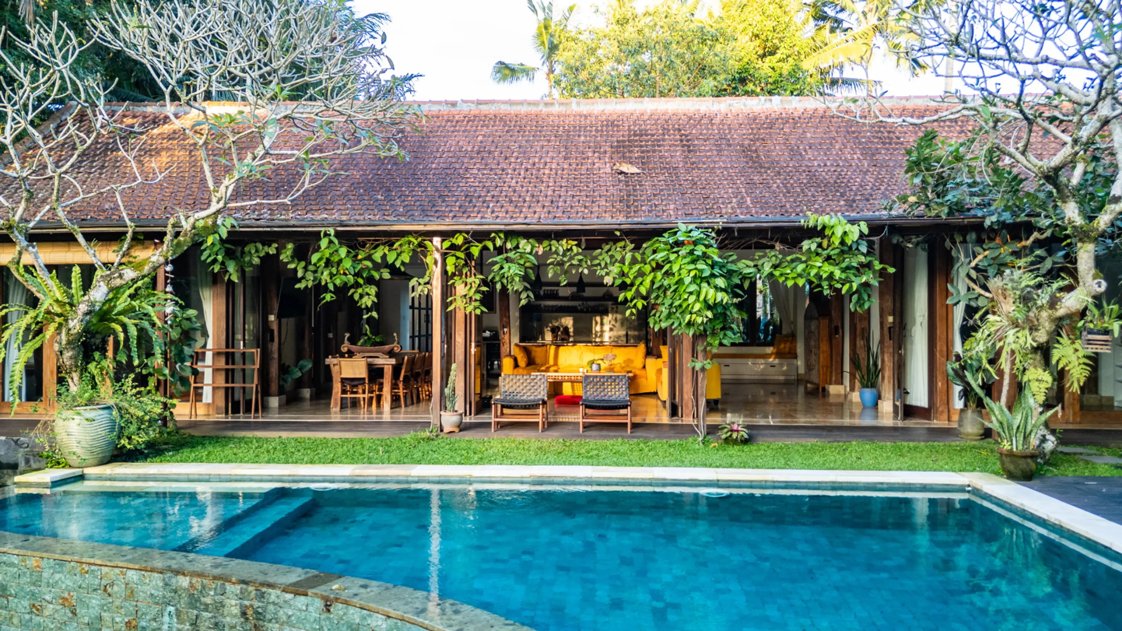 A swimming pool in front of a tropical villa with an open-air living area.
