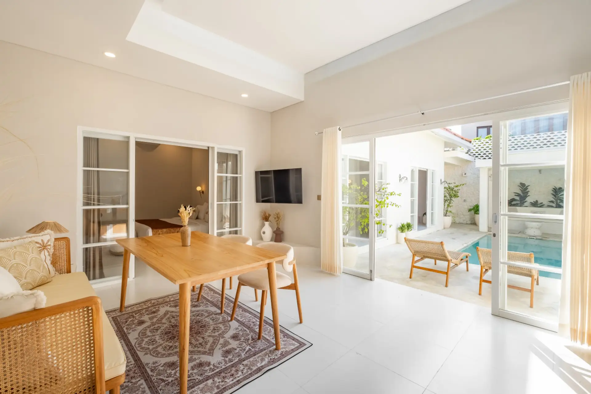 A bright and modern open-plan living space featuring a dining area with a wooden table, bench, and chairs, a wall-mounted TV, and large glass doors leading to an outdoor area.