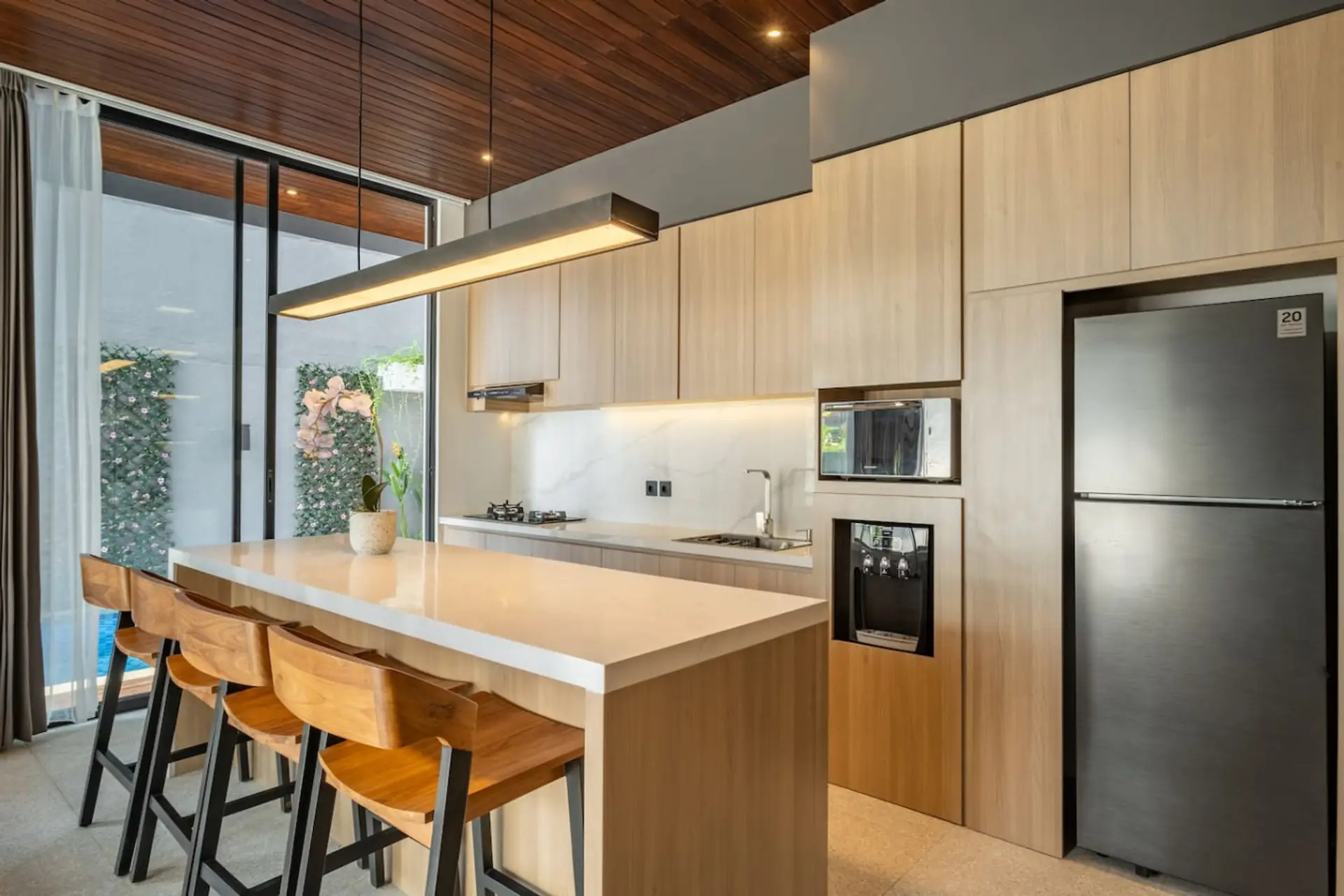 A modern kitchen with light wood cabinetry, a sleek island with bar stools, built-in appliances, and large glass doors providing natural light.