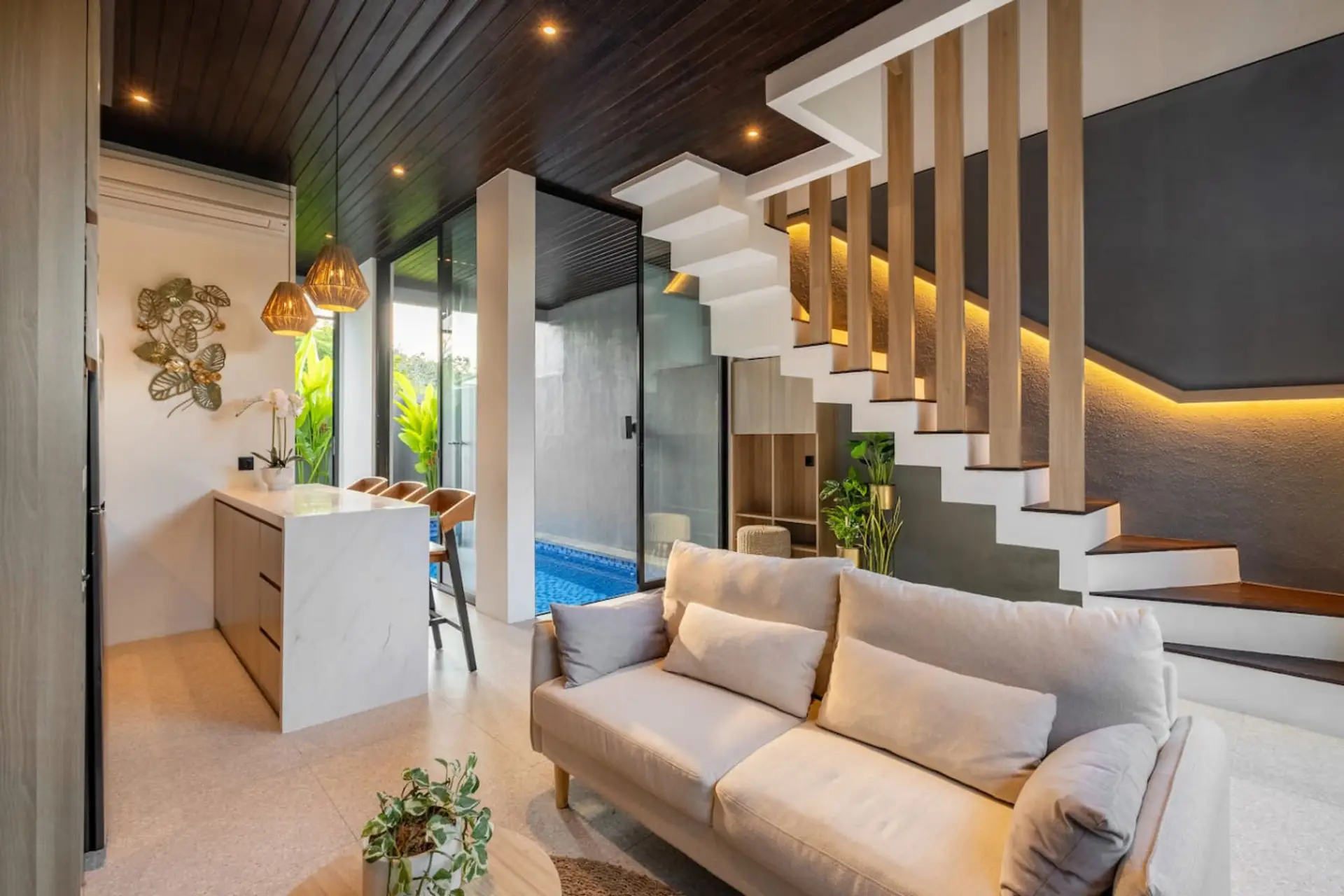 A bright, modern open-plan living area with a cozy sofa, stylish kitchen with bar seating, and large glass doors overlooking a private pool.