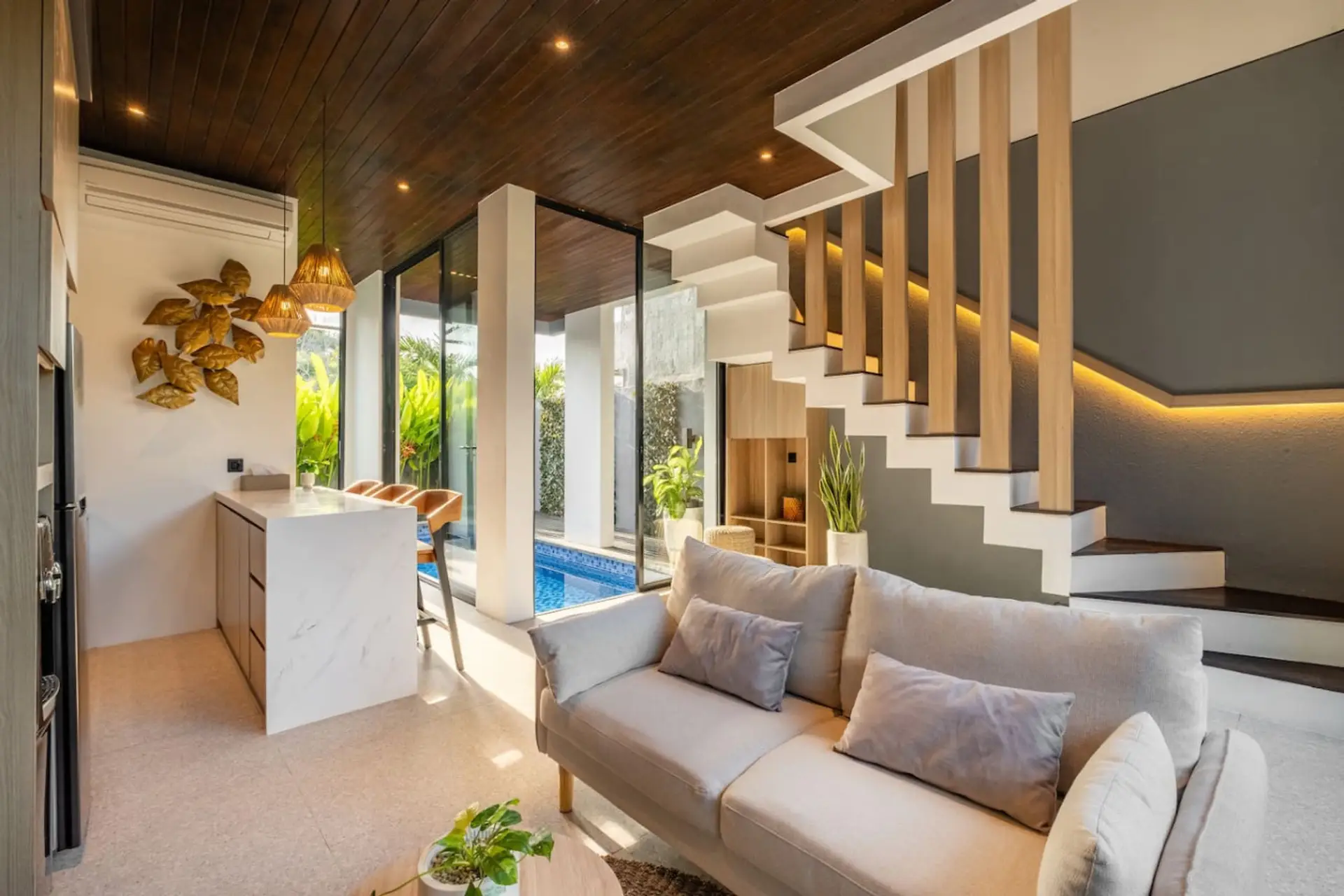 A bright, modern open-plan living area with a cozy sofa, stylish kitchen with bar seating, and large glass doors overlooking a private pool.