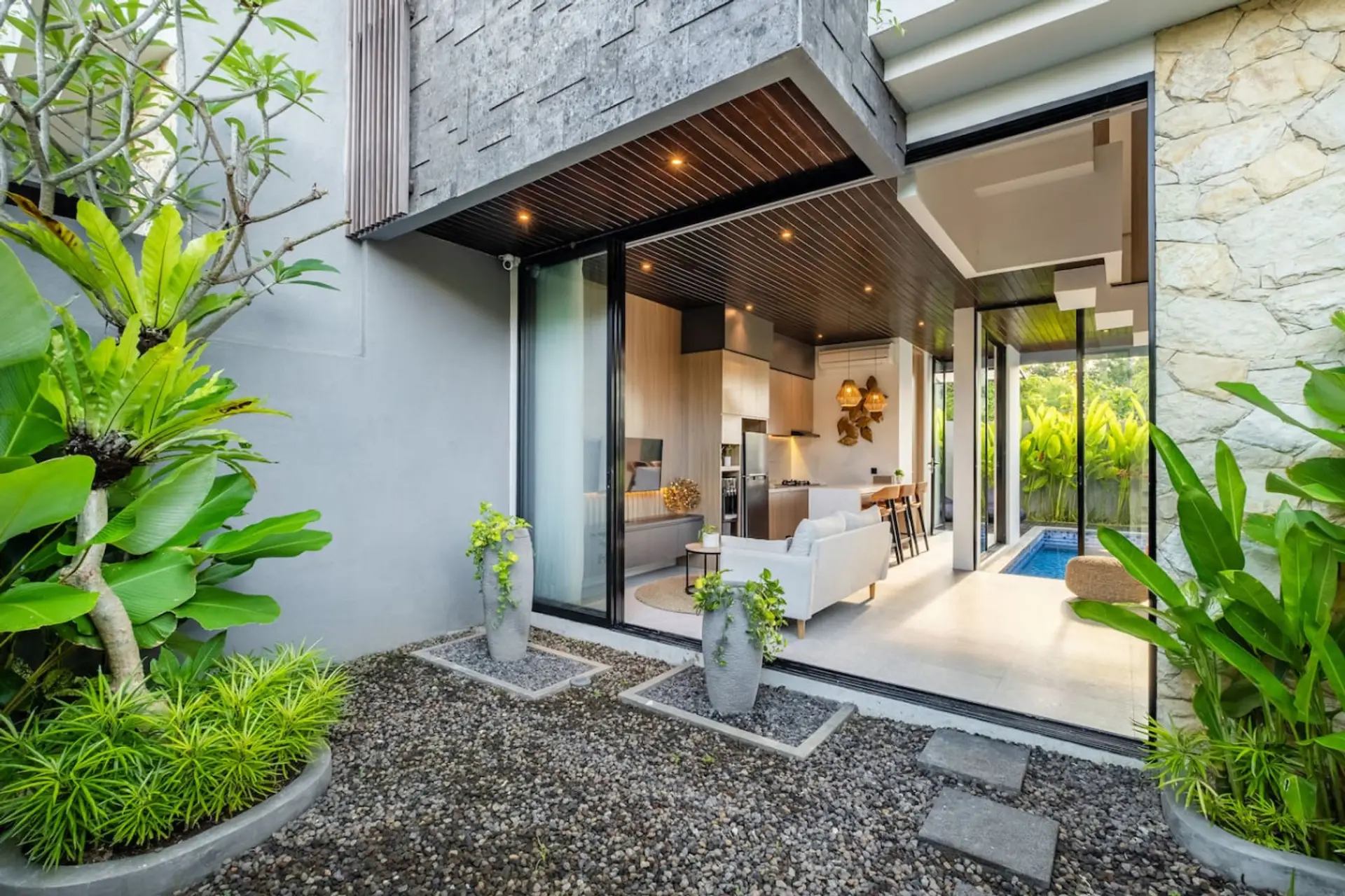 A bright, modern open-plan living area with a cozy sofa, stylish kitchen with bar seating, and large glass doors overlooking a private pool.