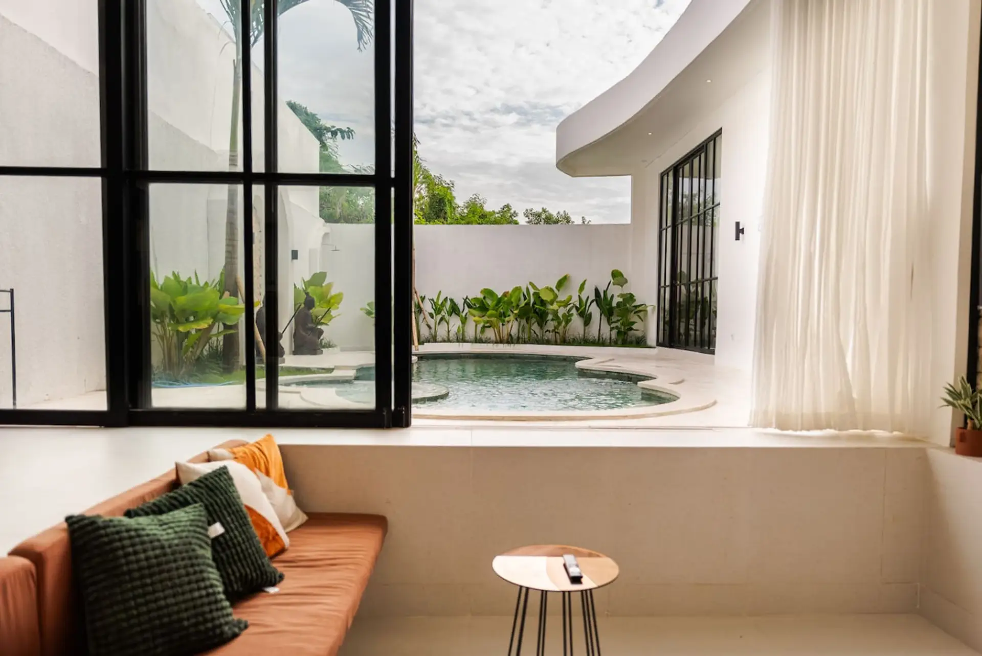 Inviting lounge space with a cozy modern sofa and vibrant cushions, offering a panoramic view of the tropical courtyard and curved pool through expansive black-framed glass windows and sheer curtains.