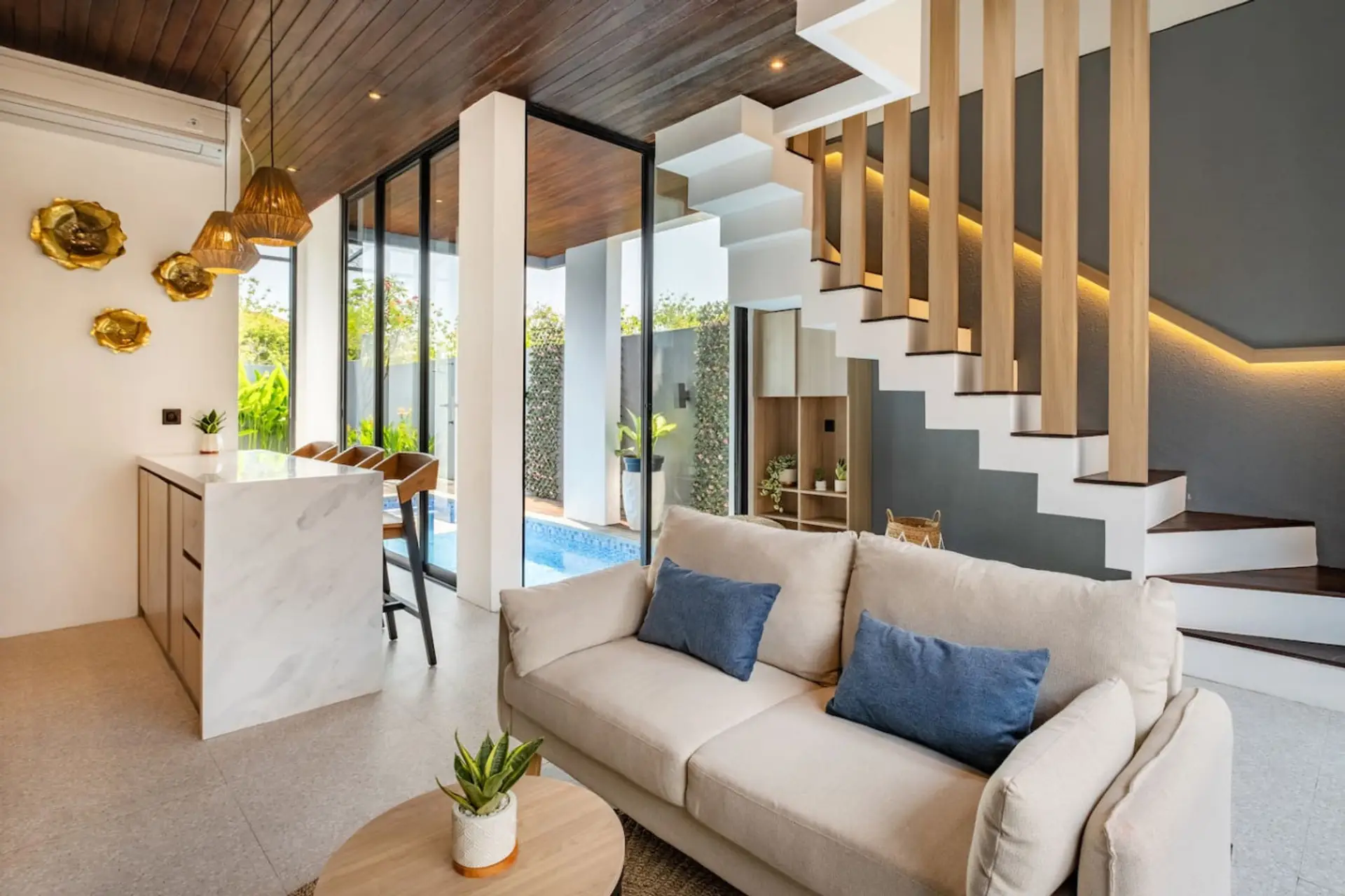 A bright, modern open-plan living area with a cozy sofa, stylish kitchen with bar seating, and large glass doors overlooking a private pool.