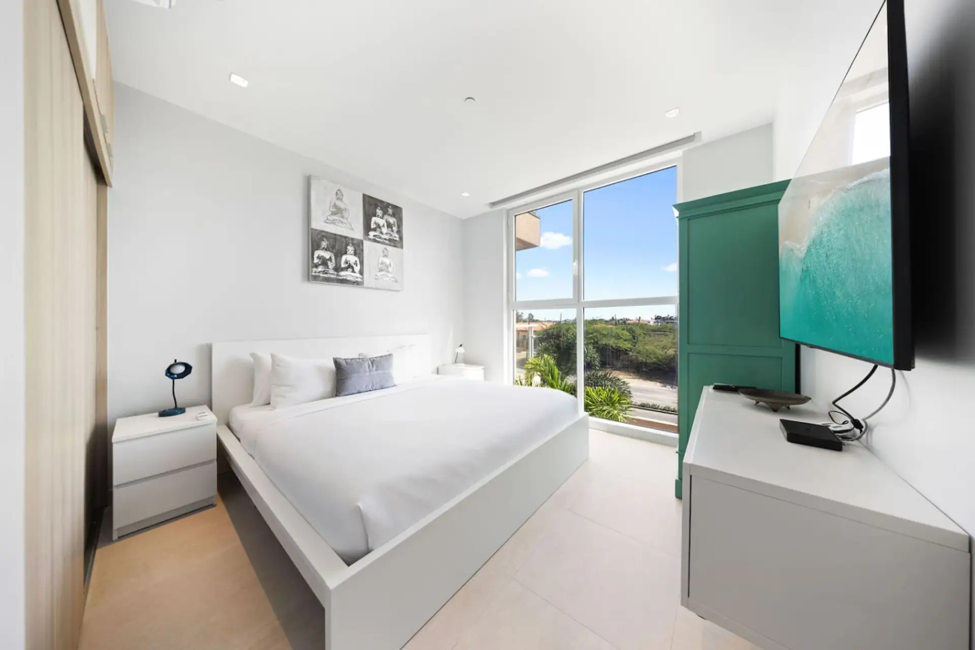 Clean, modern bedroom with a white platform bed and nightstands. A large window offers a view of the outdoors, and a turquoise cabinet adds a pop of color. The room has light tiled floors and features a set of four black-and-white photos on the wall.