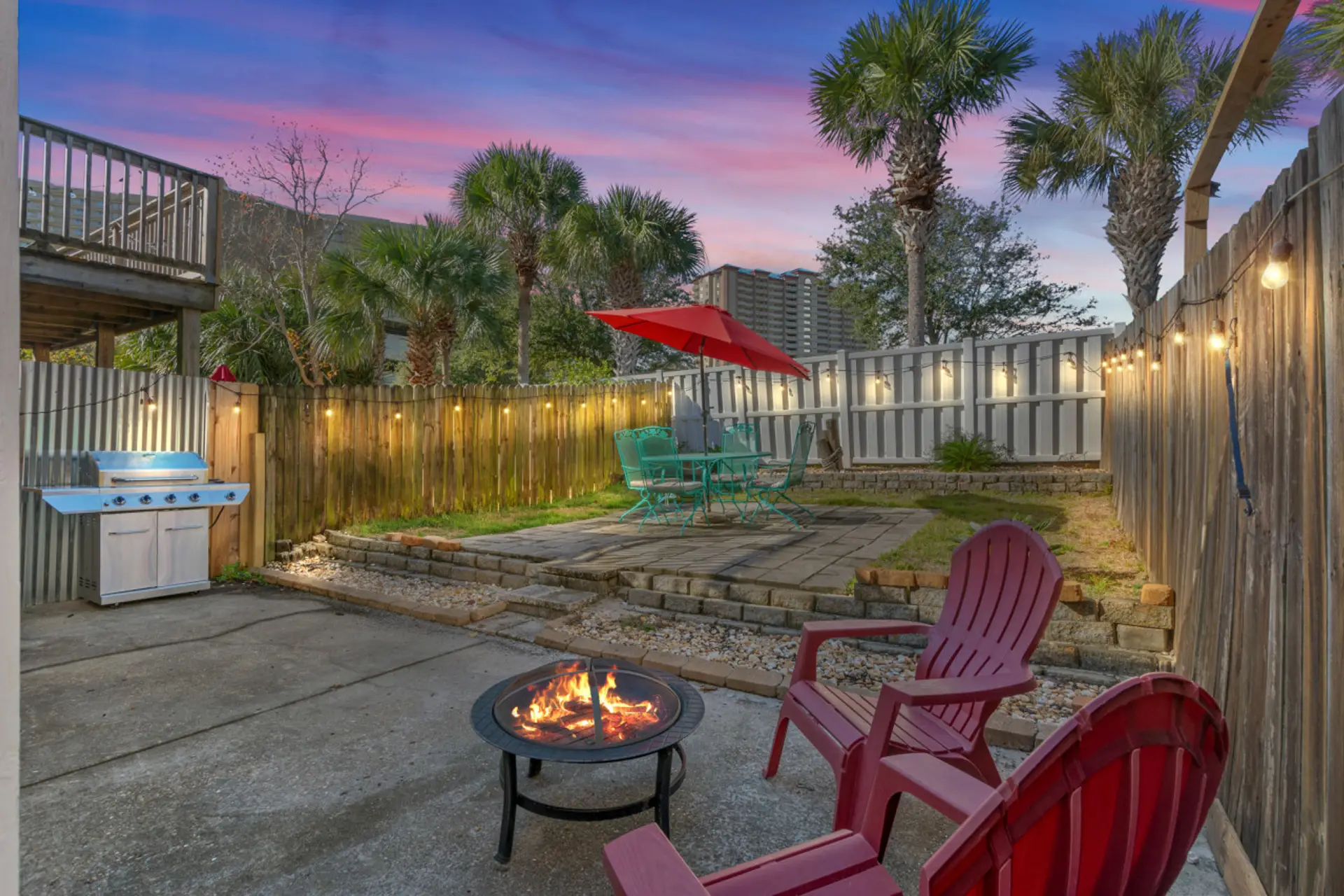 Enjoy the backyard at sunset, complete with comfortable seating under an umbrella and soft Edison lights overhead, offering a serene and inviting atmosphere for unwinding.