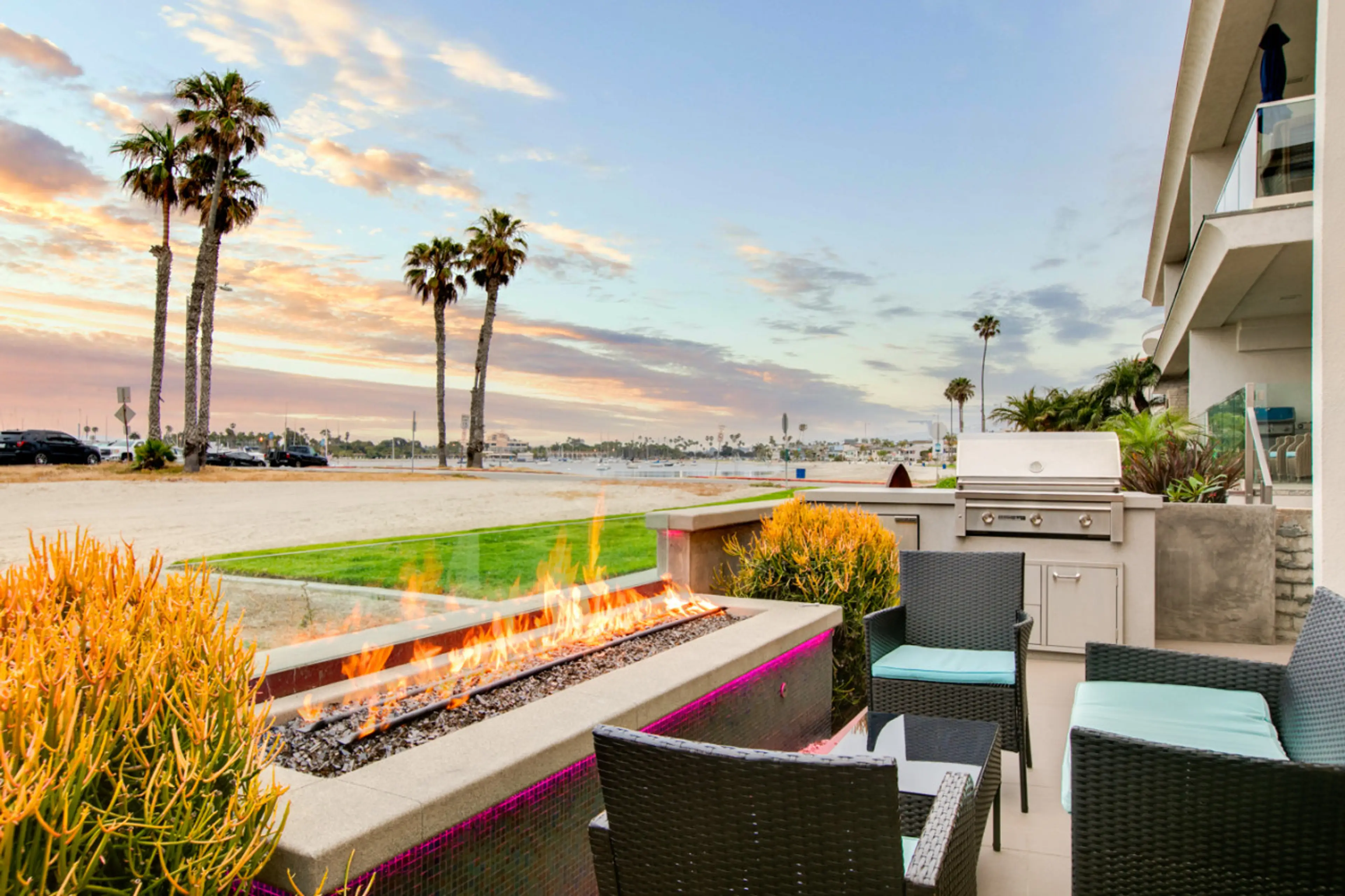 A cozy beachfront patio featuring a modern fire pit, comfortable wicker seating with blue cushions, and seamless access to the indoor living space. Surrounded by lush tropical plants, this inviting outdoor area is perfect for relaxing and enjoying warm ev